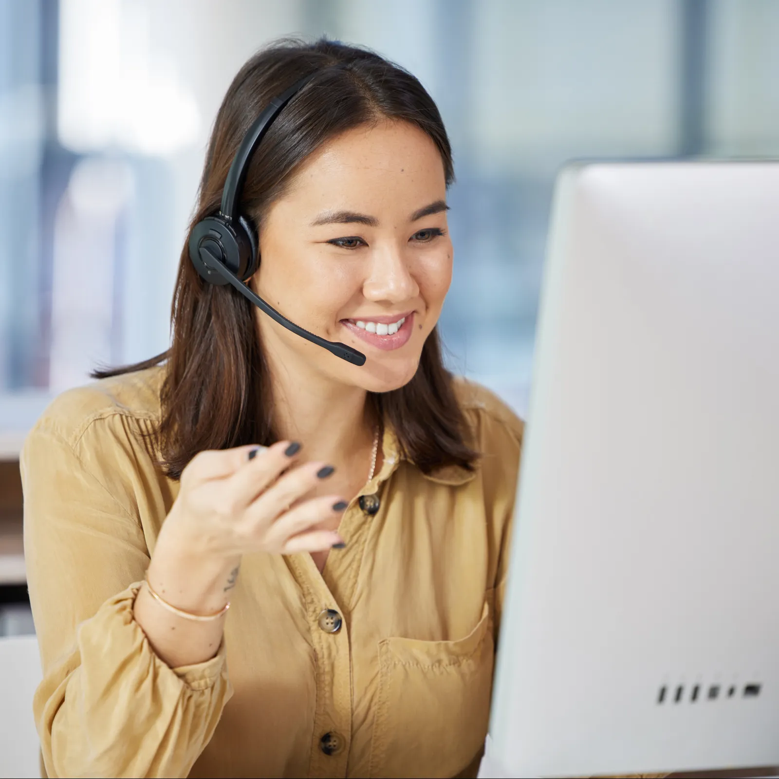 Smiling woman wearing headset and talking while looking at computer screen in a bright office.