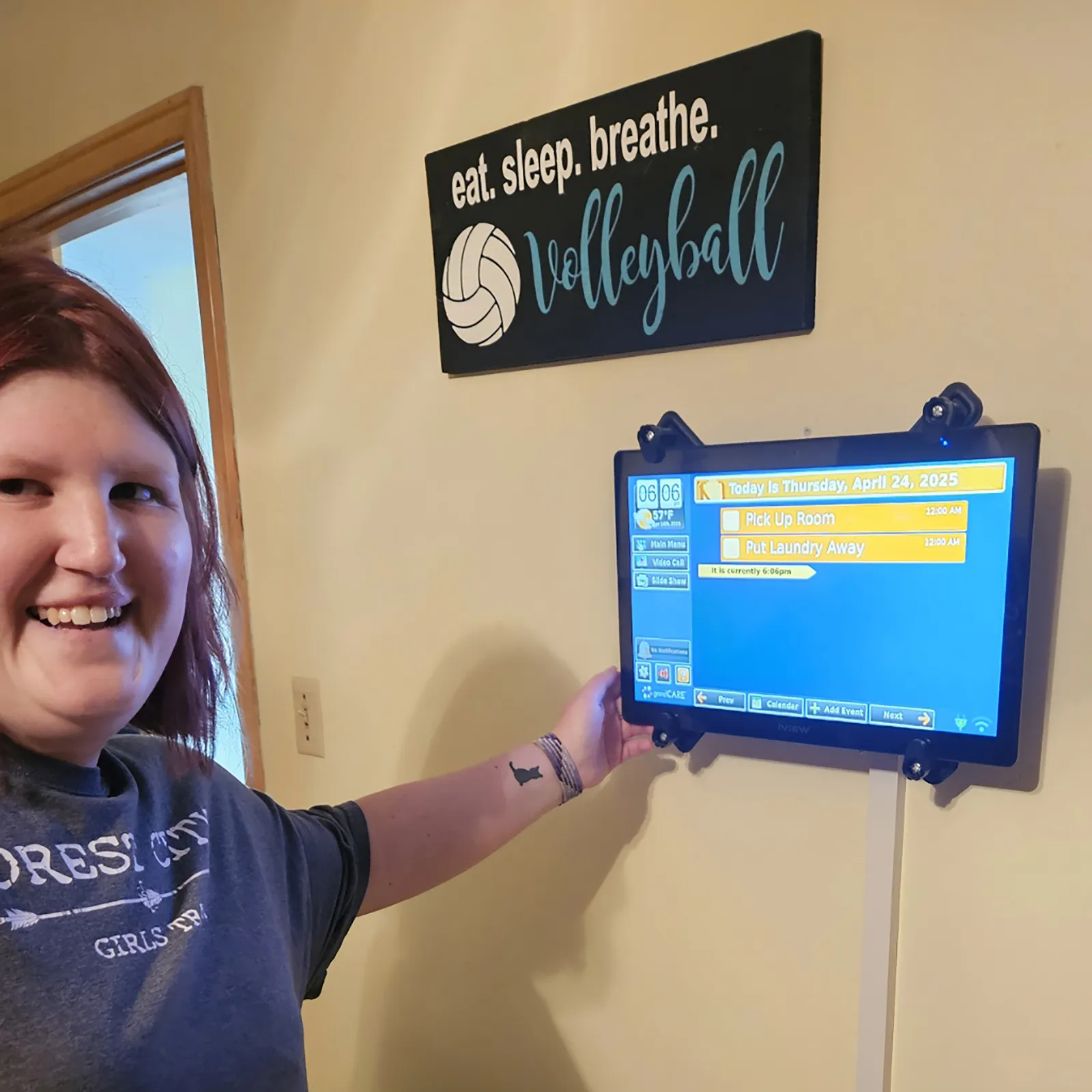 Smiling woman pointing at a wall-mounted tablet displaying a digital calendar with volleyball decor above.