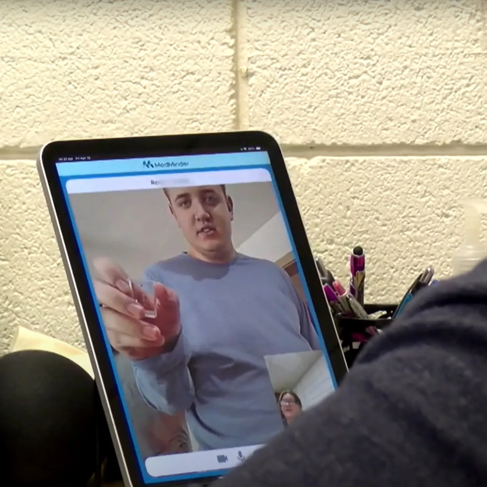 Person participating in a video call on a tablet with a man gesturing on screen in a casual indoor setting