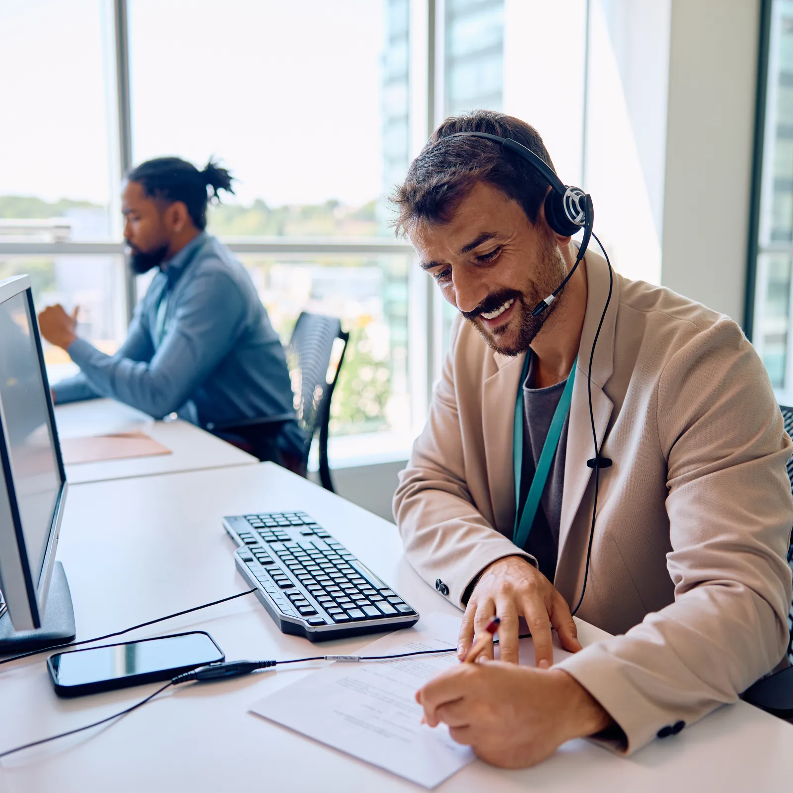 Smiling customer service agent with headset writing notes at desk with computer in modern office environment