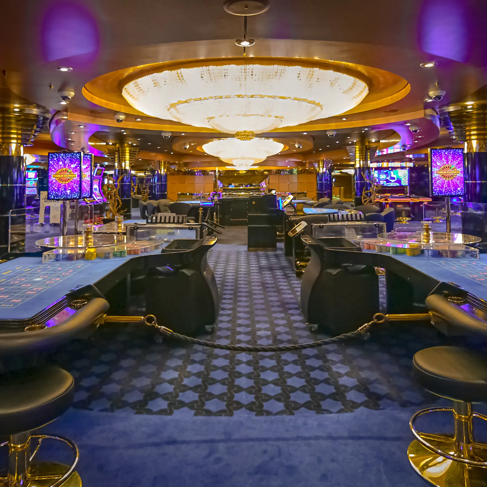 Luxurious casino interior with poker tables, slot machines, blue carpet, and golden chandeliers under purple lighting.