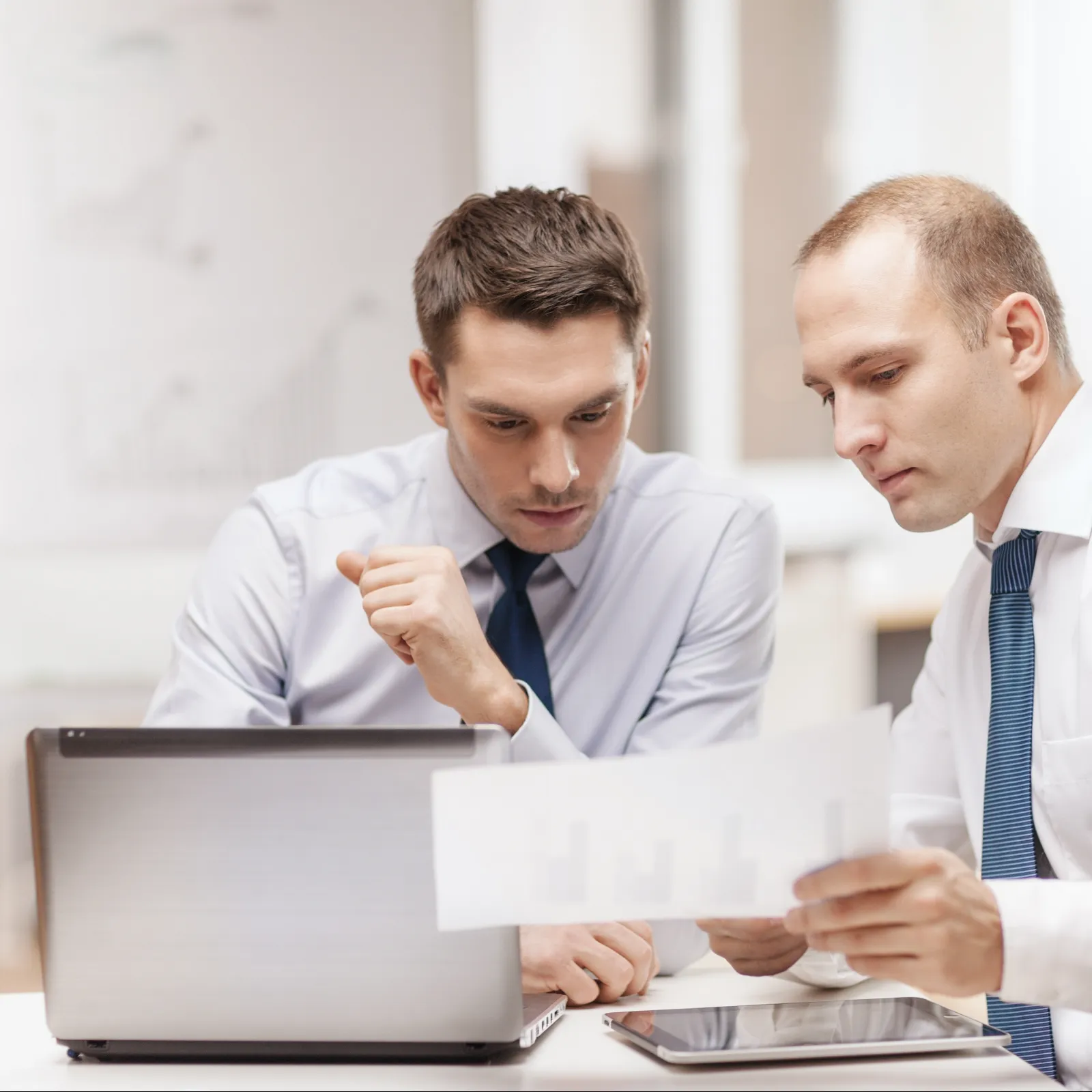 Two businessmen in white shirts and ties review documents and laptop data in a modern office setting.