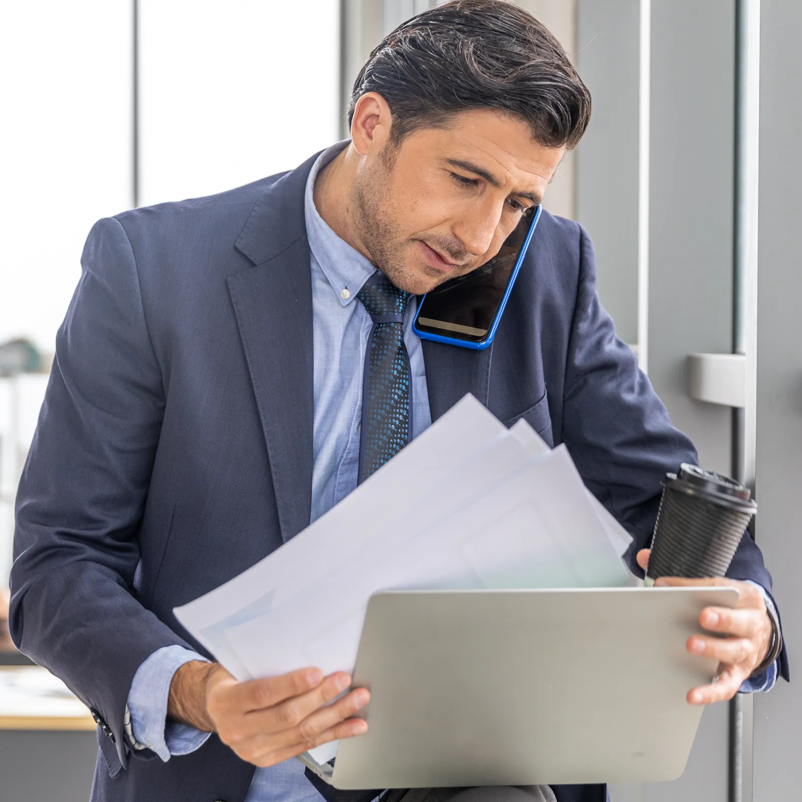 Businessman multitasking with phone, papers, laptop, and coffee in modern office setting.