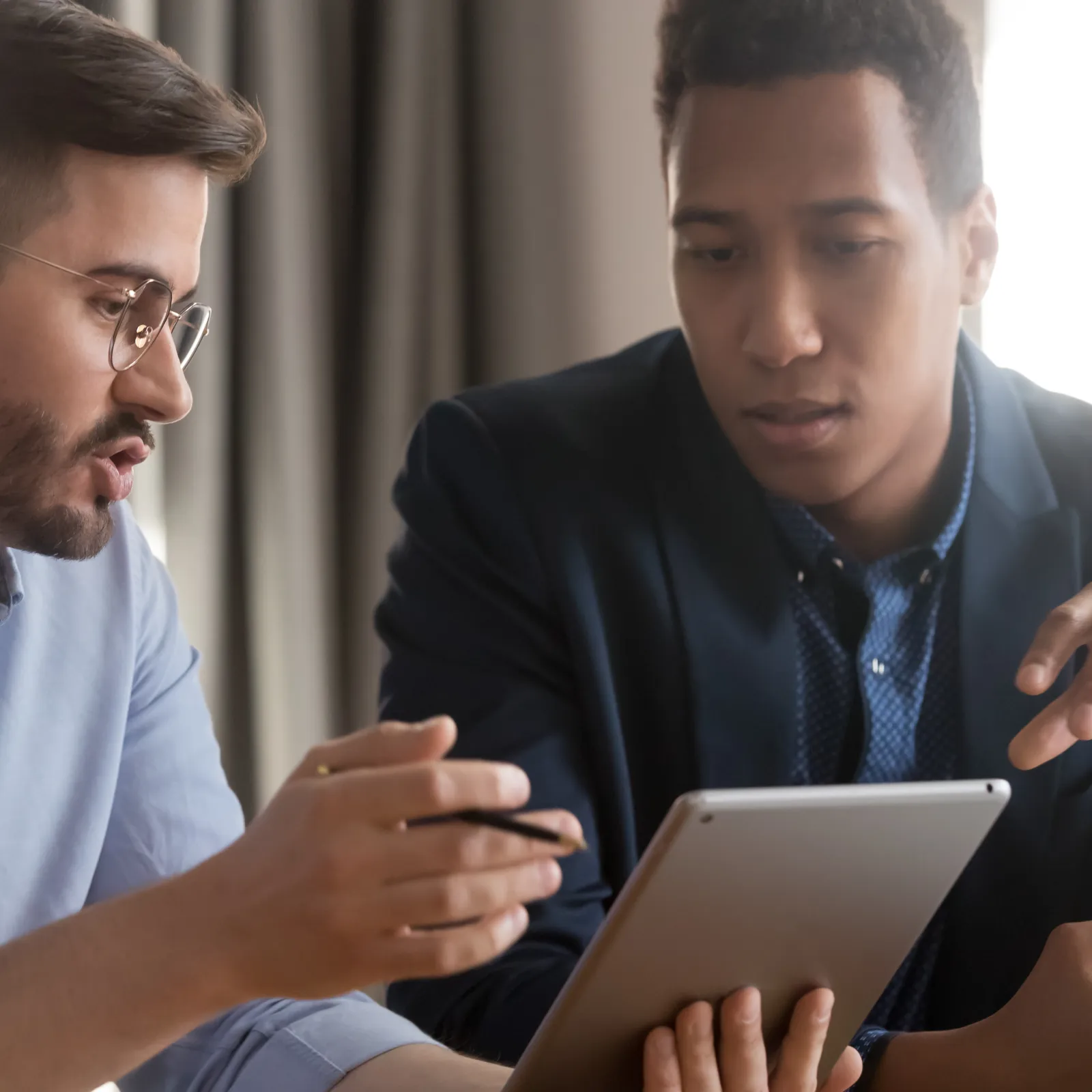 Two diverse men in business attire discussing information displayed on a tablet during a meeting.