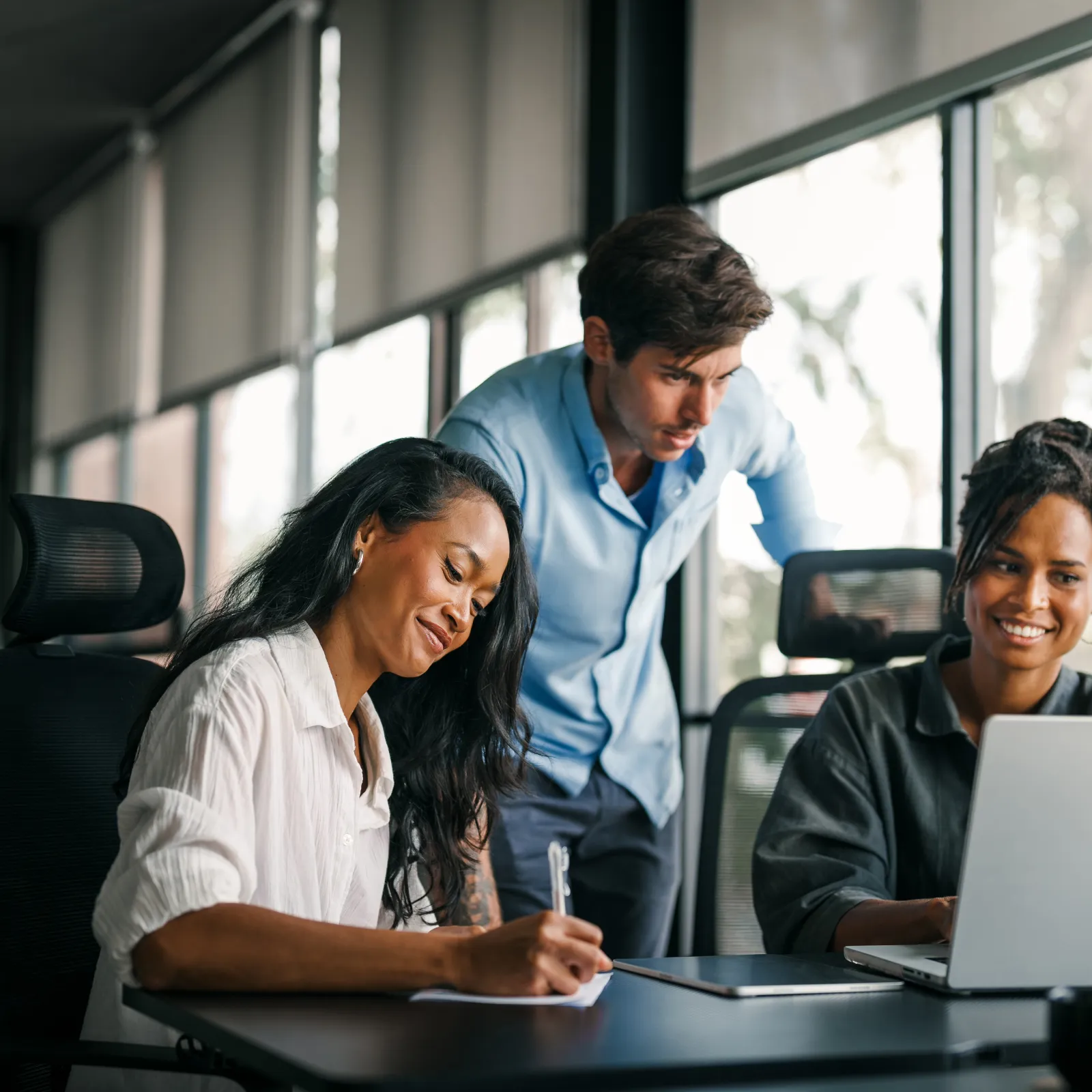 Three diverse colleagues collaborate happily in bright modern office with laptop and notes on table