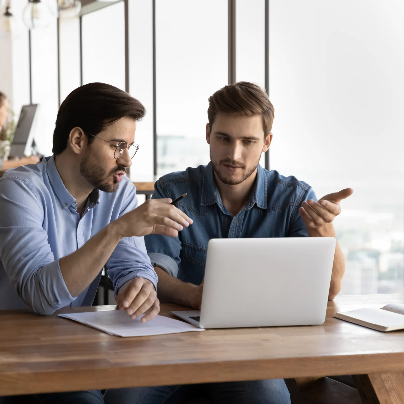 Two men discussing work on a laptop at a wooden table in a modern office with a woman in the background.