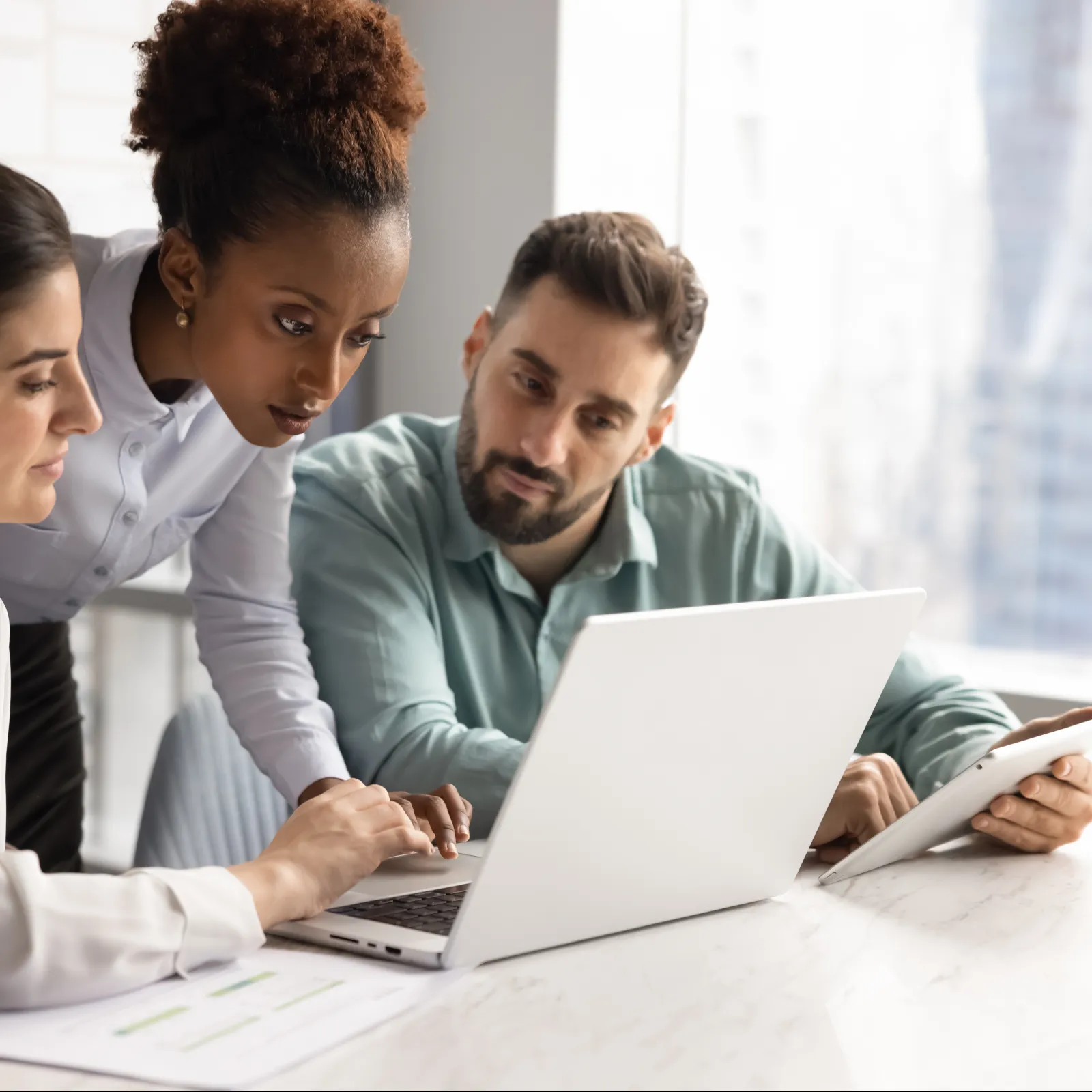 Three diverse colleagues collaborating over a laptop in a modern office with city views, discussing work intently