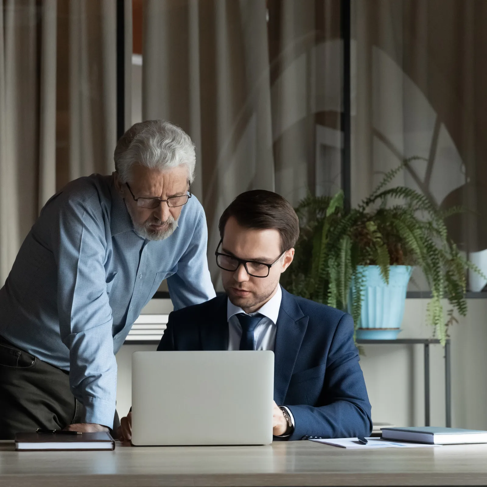 Older man mentoring younger man in suit working together on laptop in modern office setting