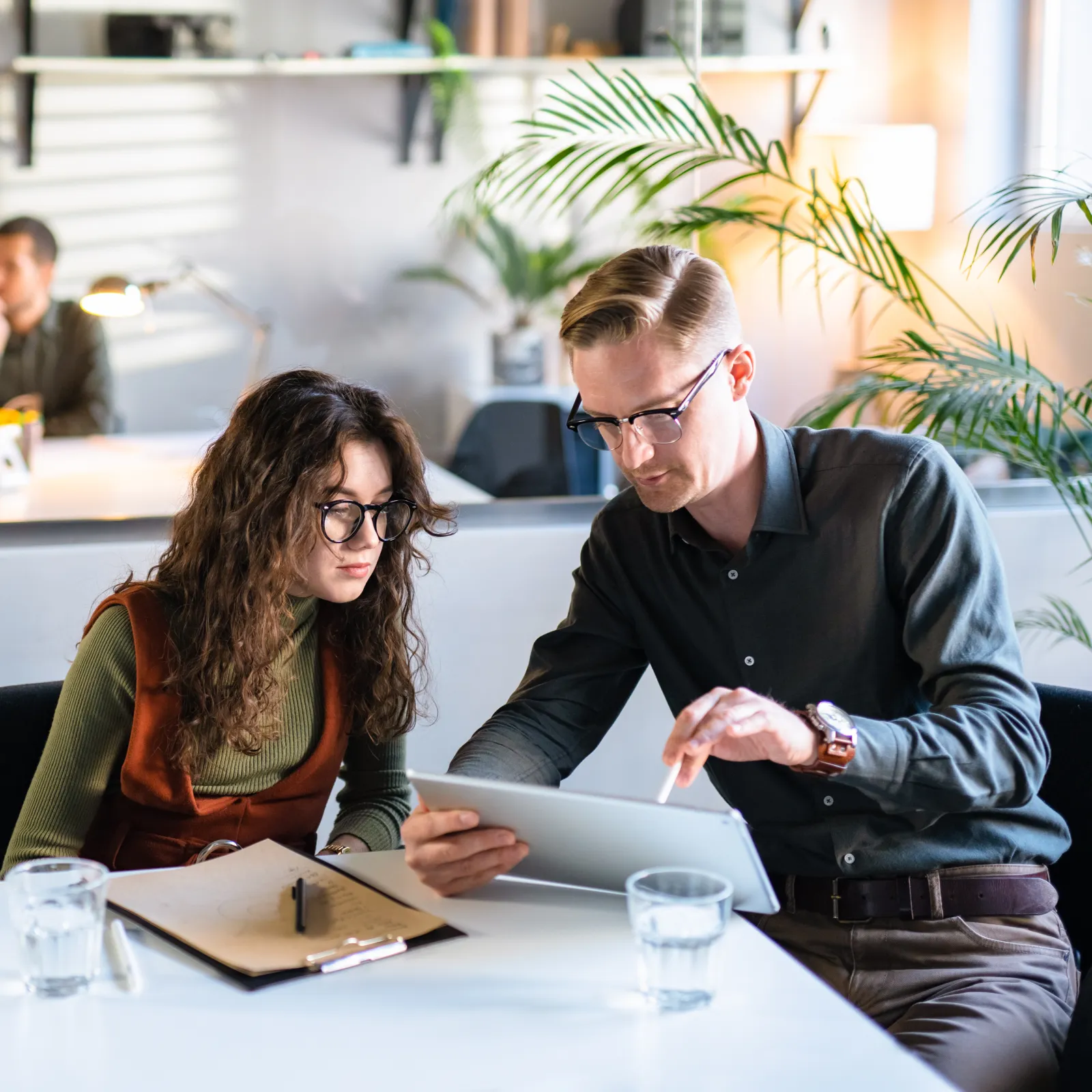 Two colleagues collaborate using a tablet in a bright modern office with plants and glass partitions.