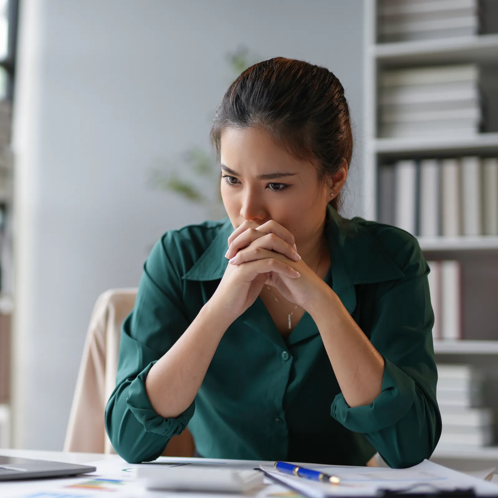Woman in green shirt looking thoughtfully at a laptop with papers and pen on desk in a modern office.