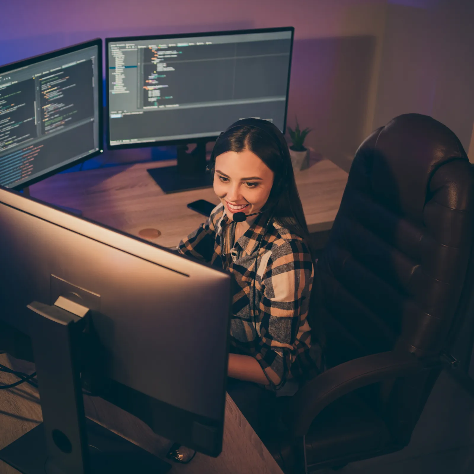 Smiling woman with headset working on multiple monitors displaying code in a dimly lit modern office space.