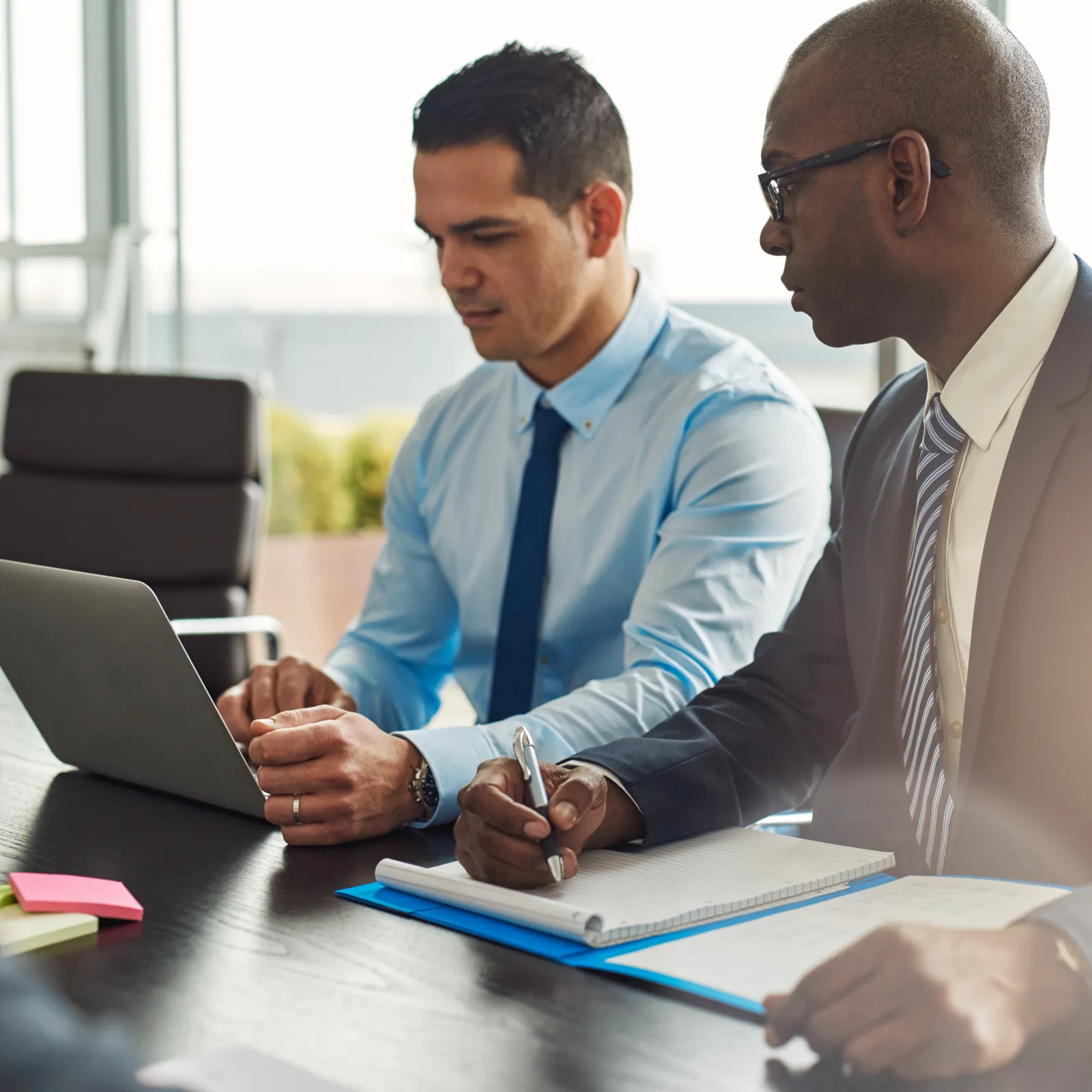Two business professionals in suits collaborating with laptop and notes during a meeting in a bright office.