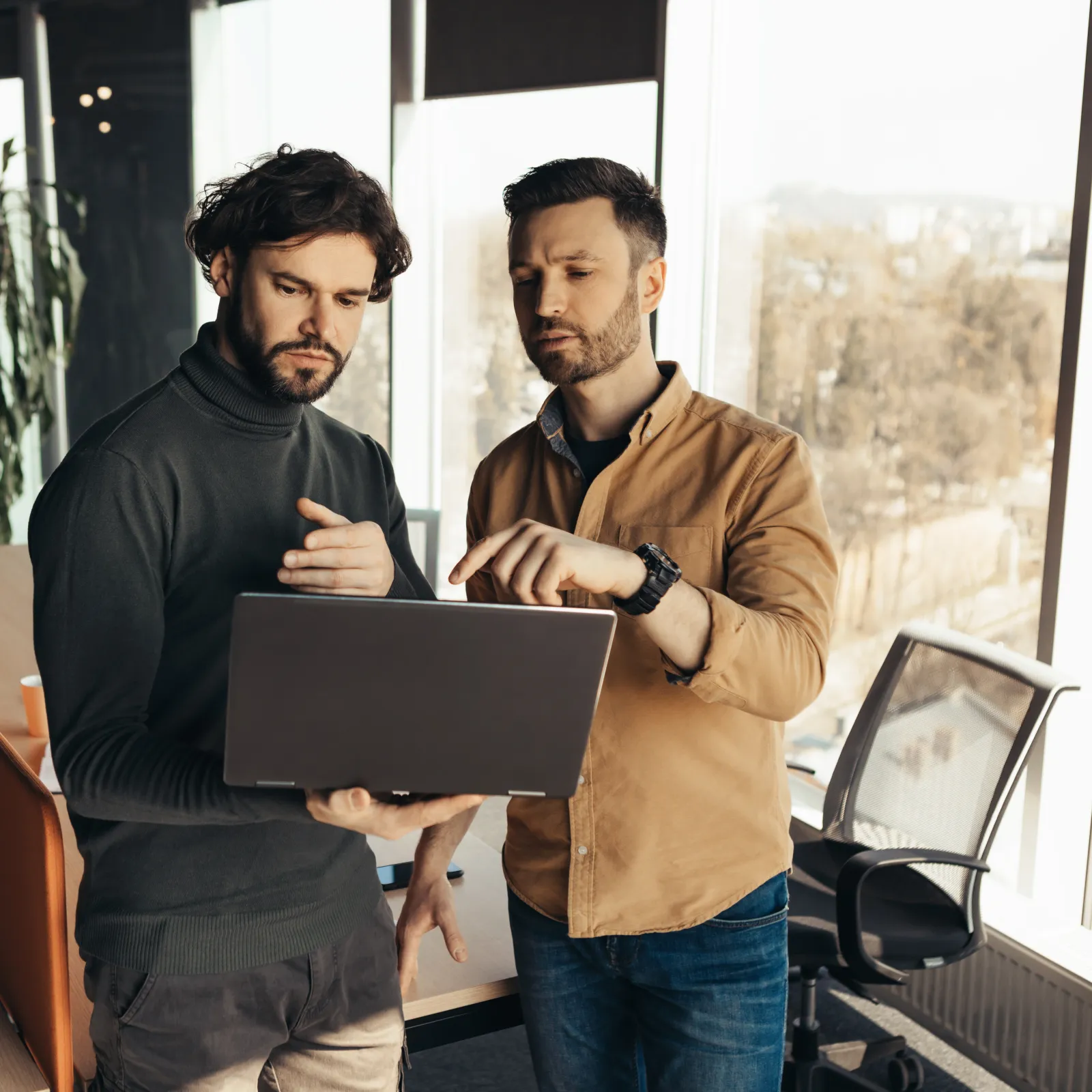 Two men discussing work while viewing content on a laptop in a modern office with large windows.
