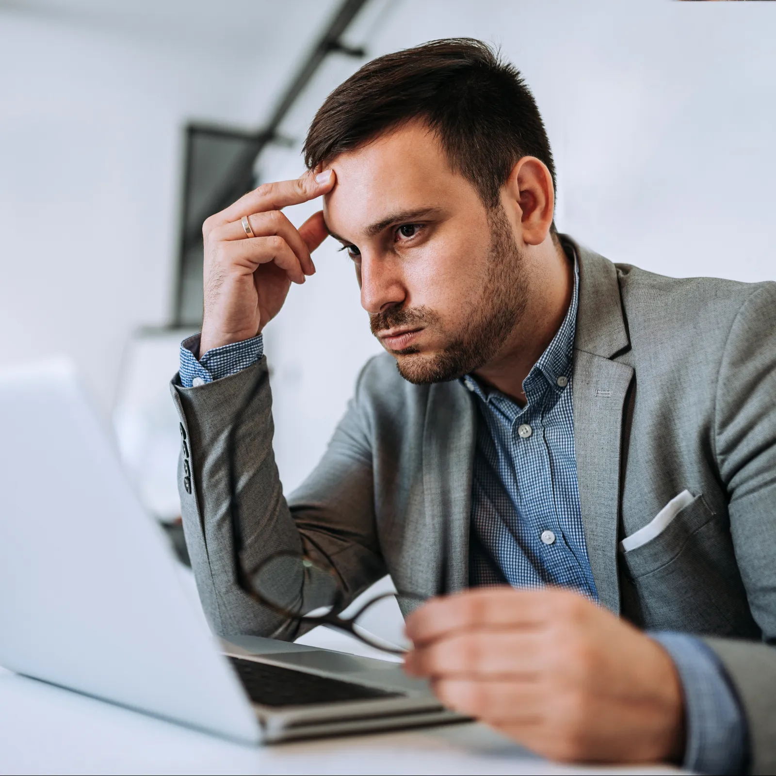 Pensive young man in gray suit working on laptop, holding glasses, focused in modern office setting.