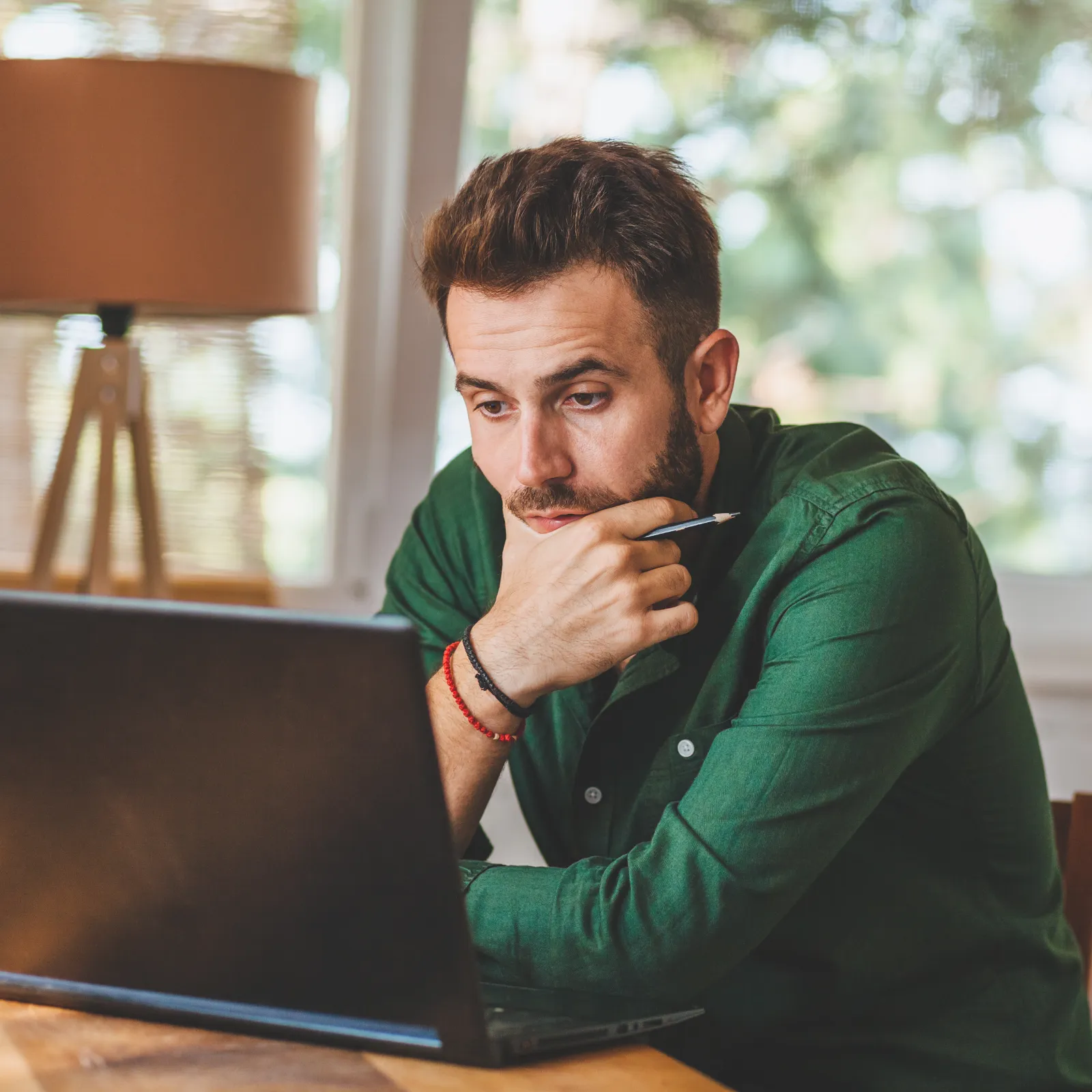 Man in green shirt working focused on laptop at home office with natural light in background