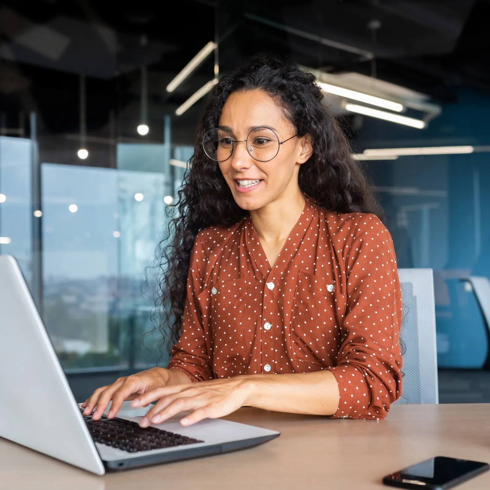 Young woman with glasses working on laptop in modern office, smiling and typing at the desk.