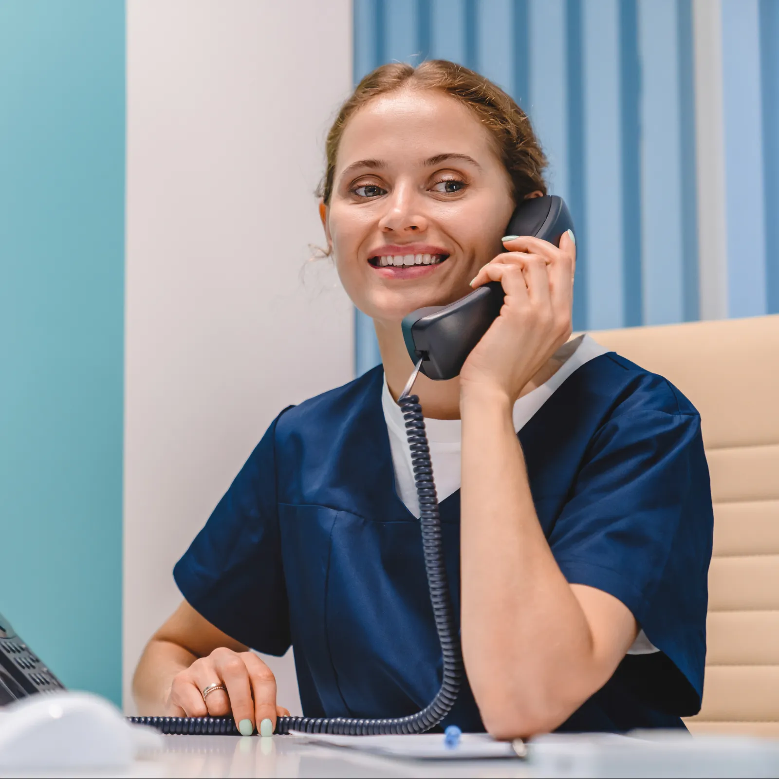 Smiling healthcare worker in navy scrubs speaking on office phone at desk with computer and paperwork