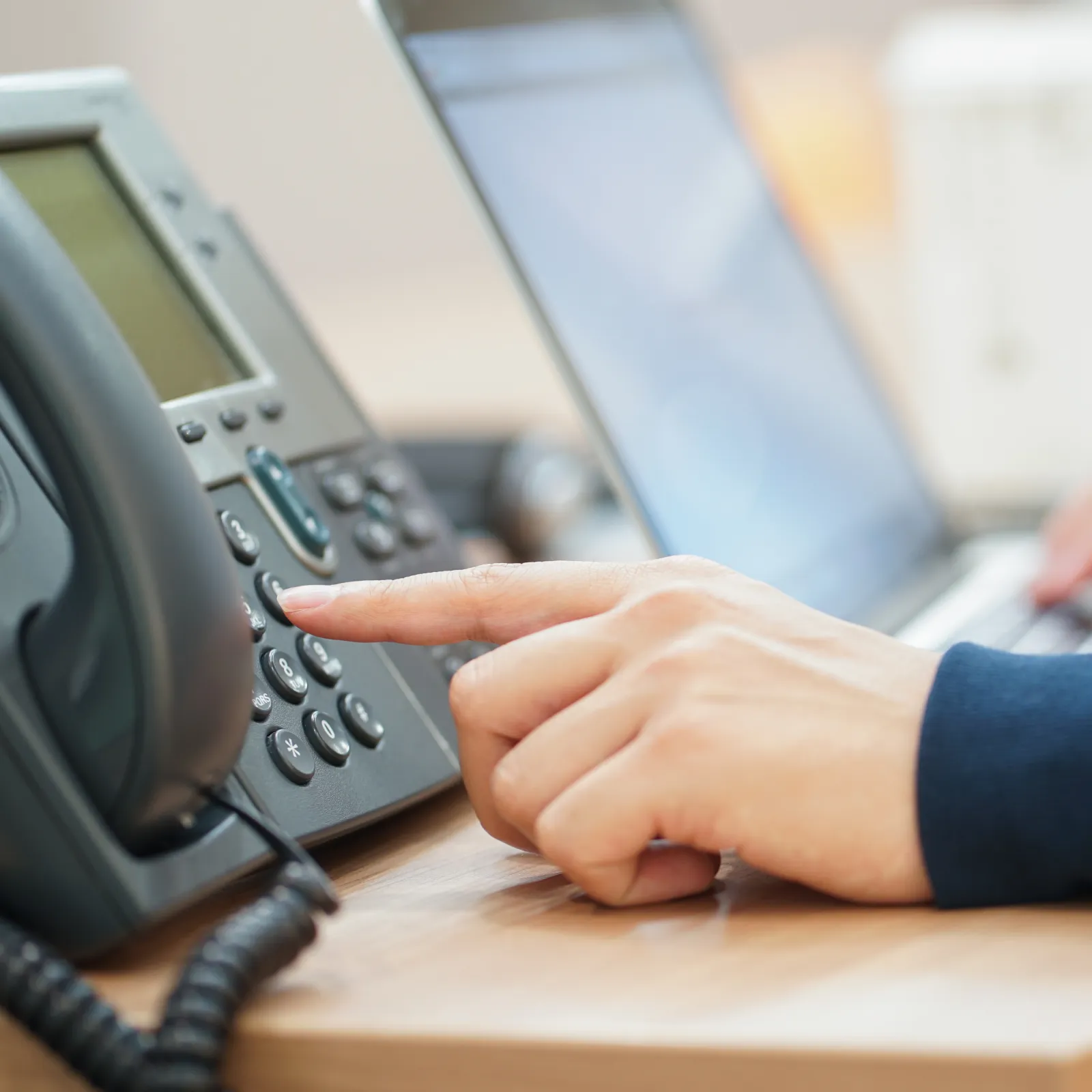 Person pressing a button on an office desk phone while typing on a laptop keyboard in the background.