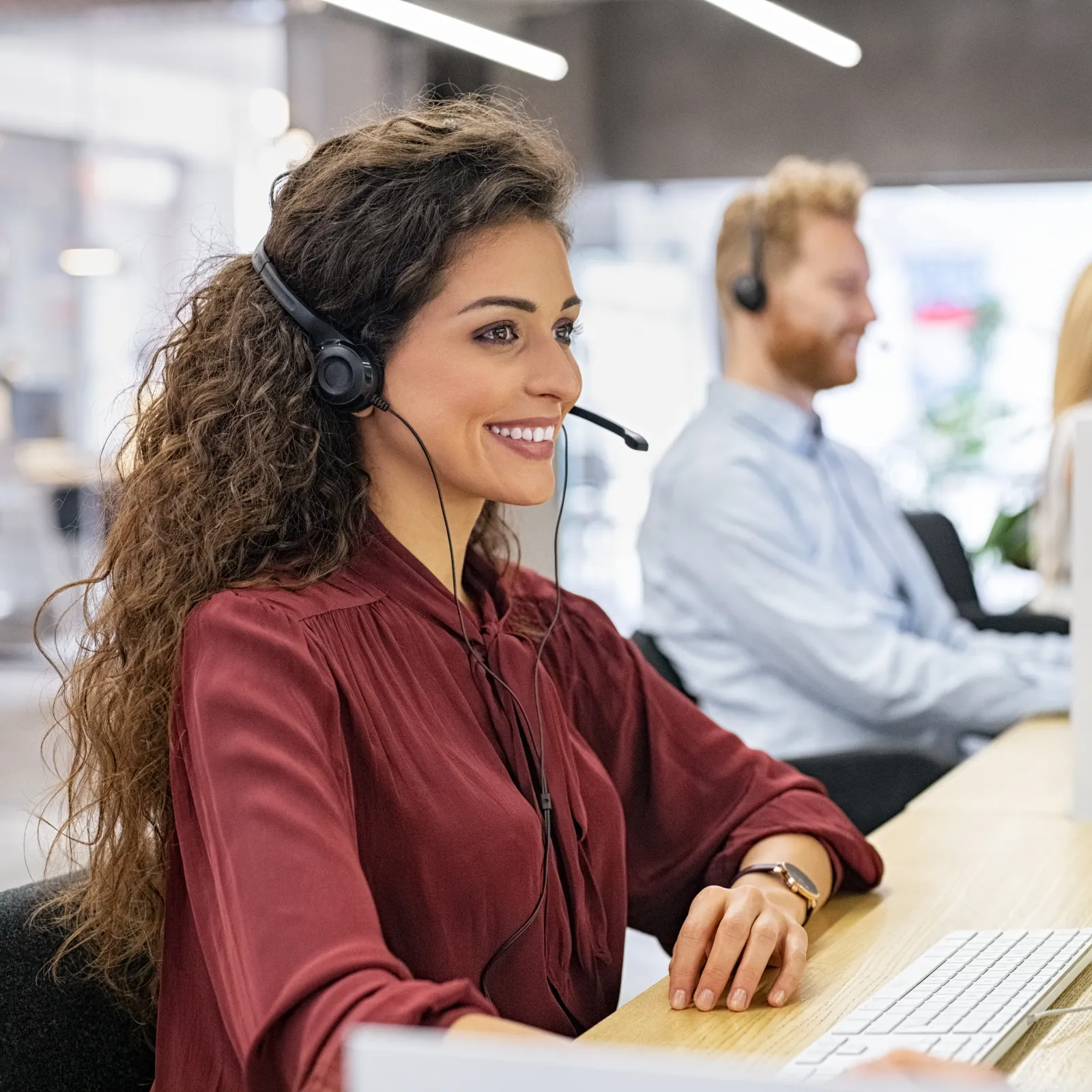 Smiling woman with headset working at computer in modern office environment with blurred background.