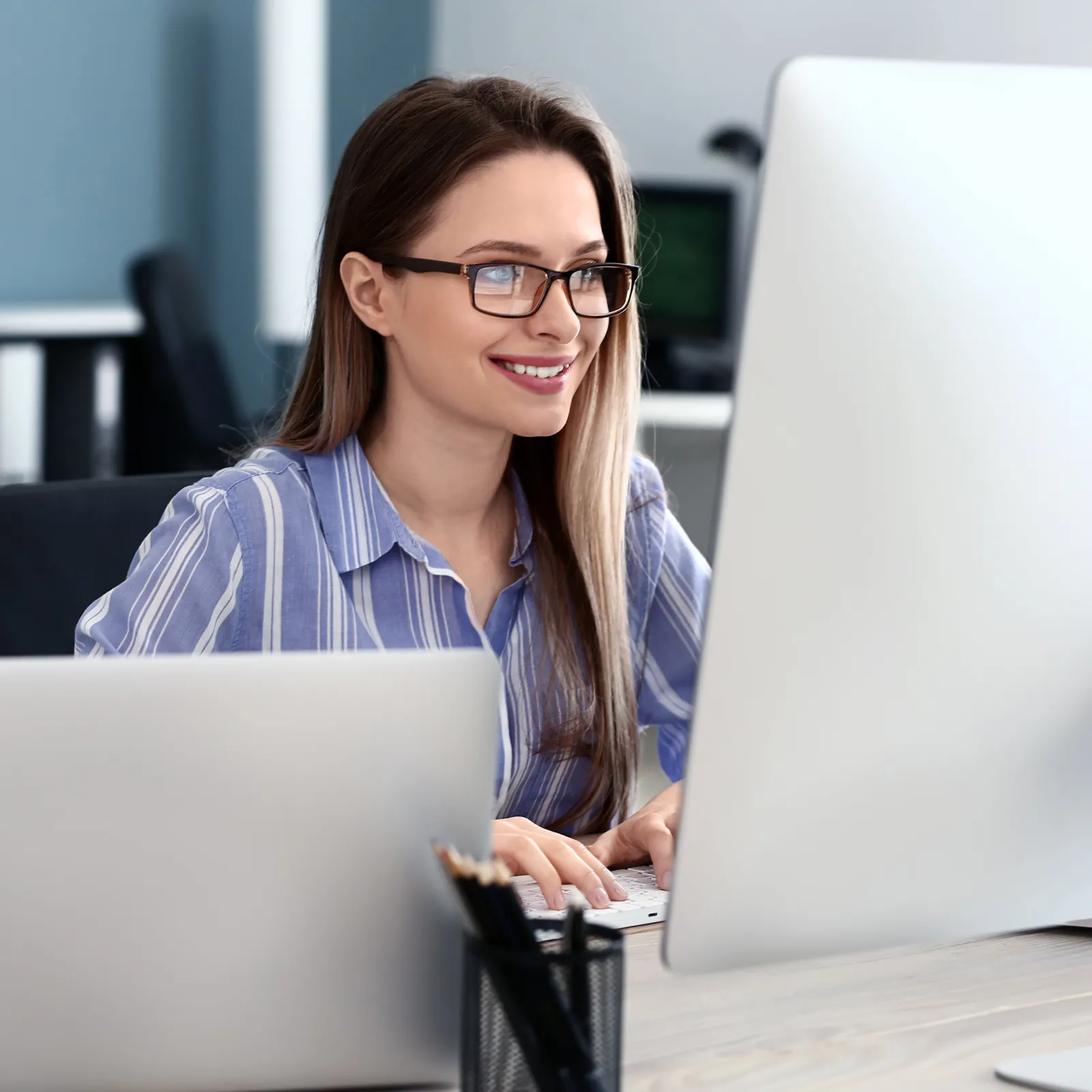 Young woman with glasses working on desktop computer in modern office smiling focused on screen