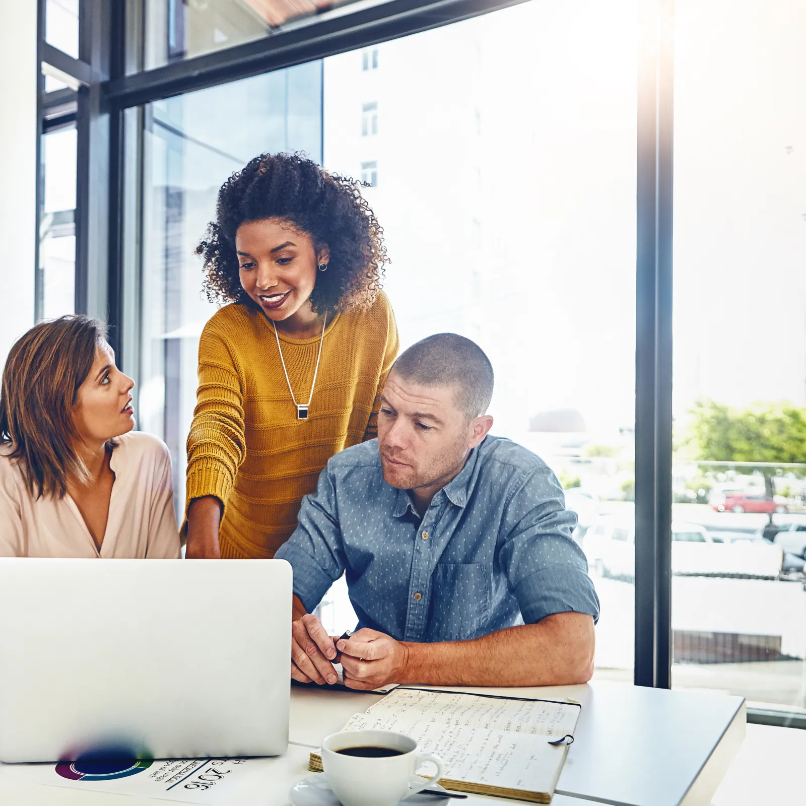 Four diverse professionals collaborating around a laptop in a modern office with large windows and natural light