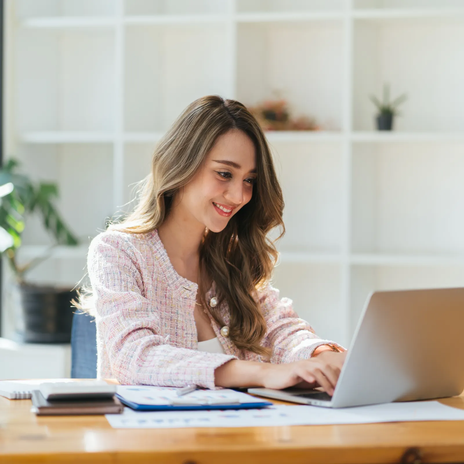 Smiling woman working on laptop at desk with papers and plants in a bright modern office space