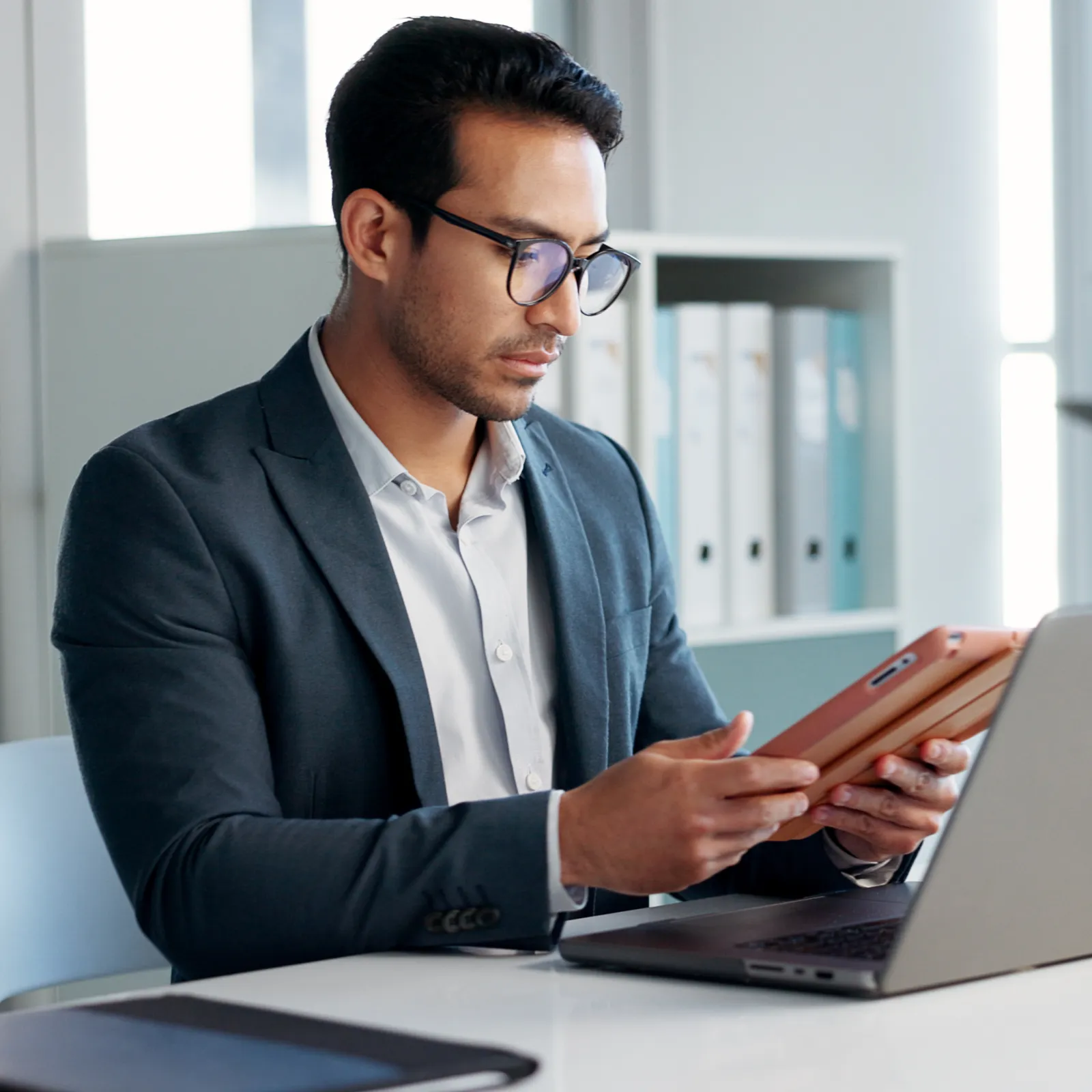 Professional man in glasses working on tablet with laptop at modern bright office desk