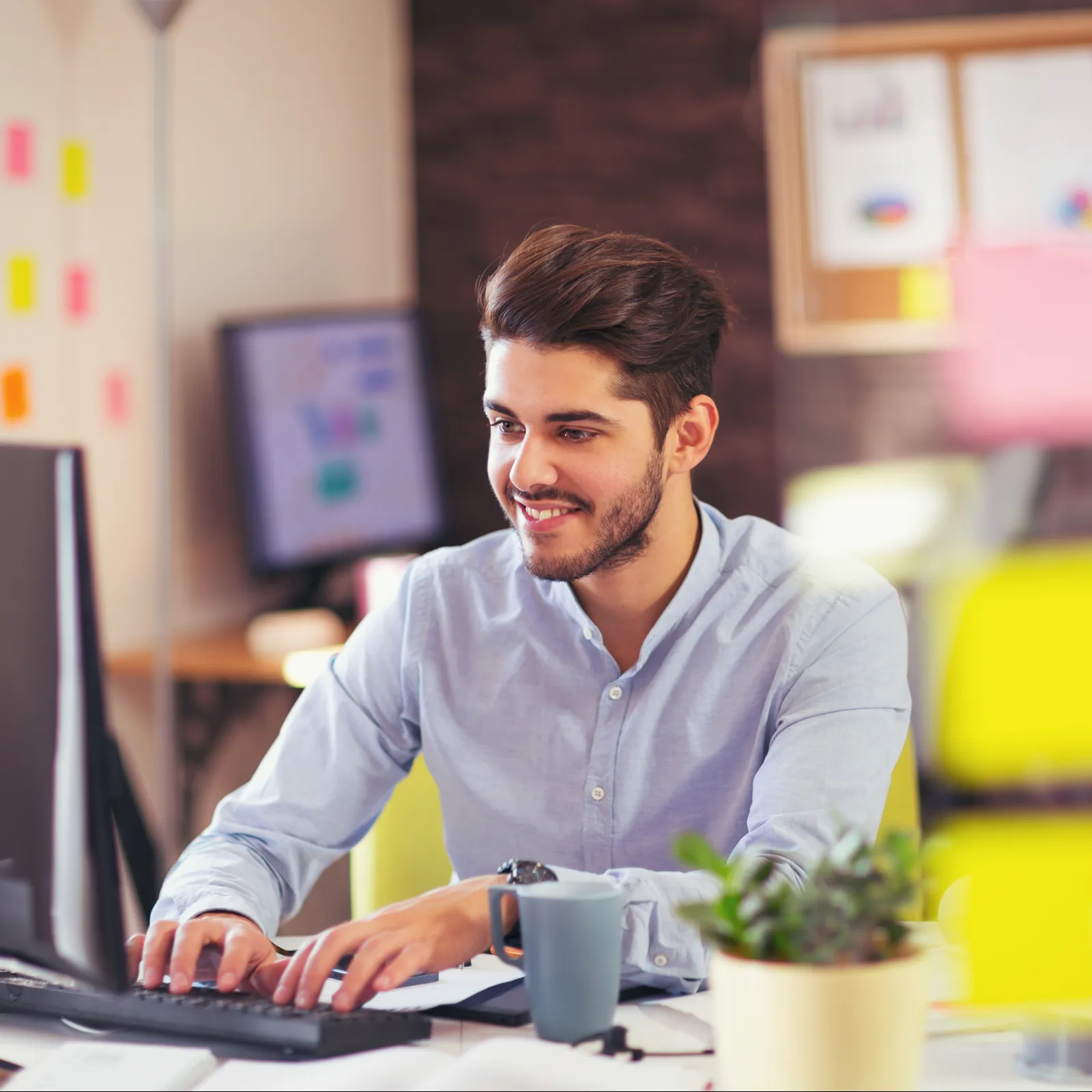 Young man working on computer in modern office with plants, coffee mug, and sticky notes around.