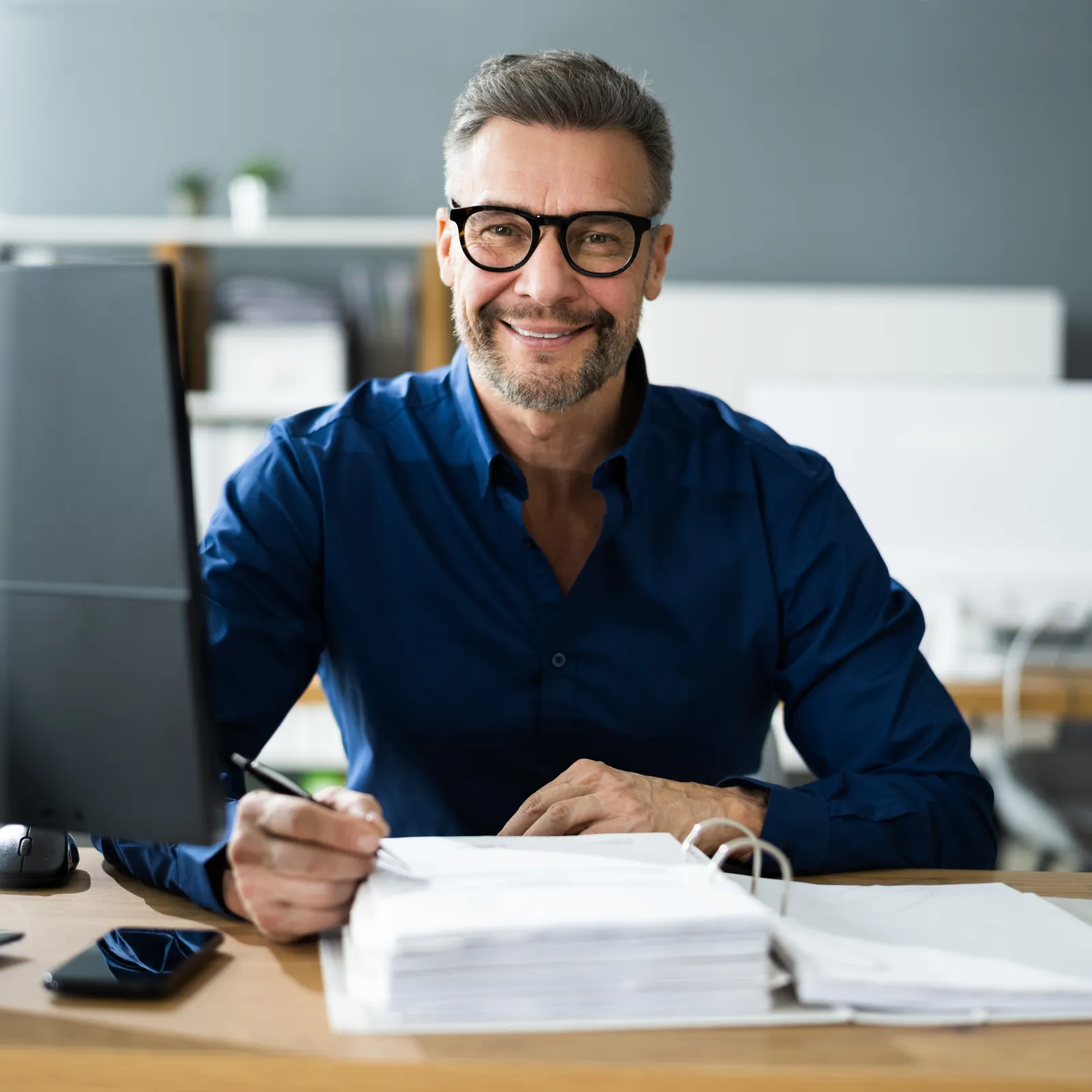 Smiling businessman with glasses working at desk surrounded by paperwork and computer in modern office