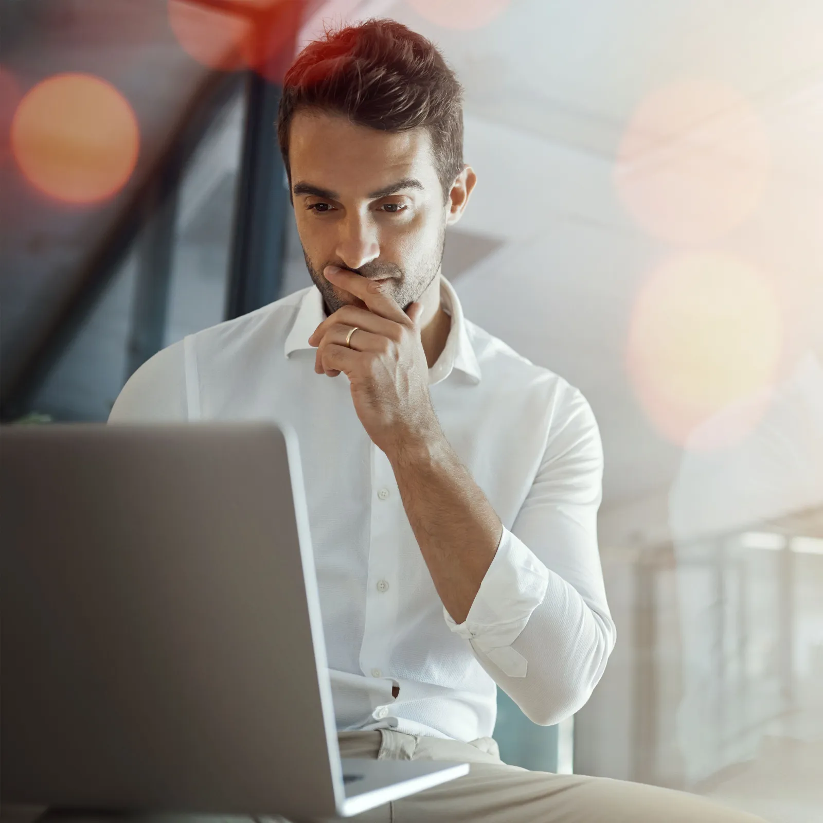 Man in white shirt thoughtfully working on laptop in modern bright office with lens flare effects.