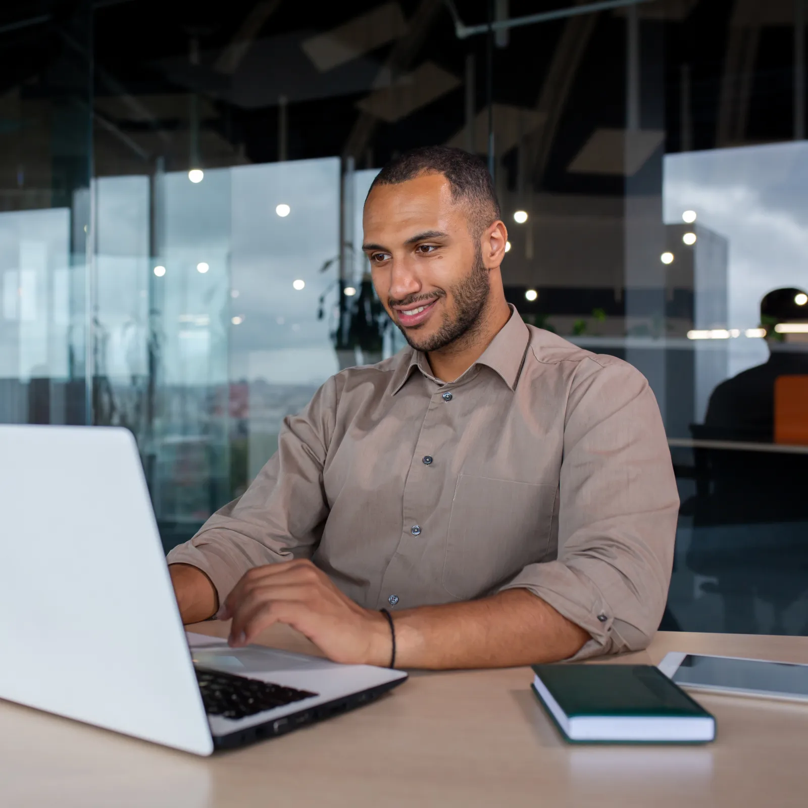 Smiling man working on a laptop in a modern office with glass walls and natural light.