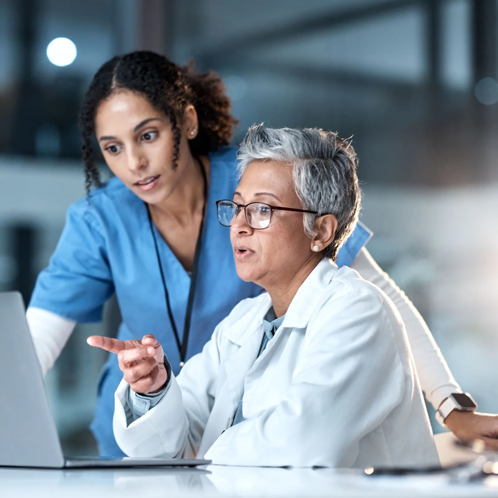 Two female medical professionals discuss patient data on a laptop in a modern healthcare facility at night