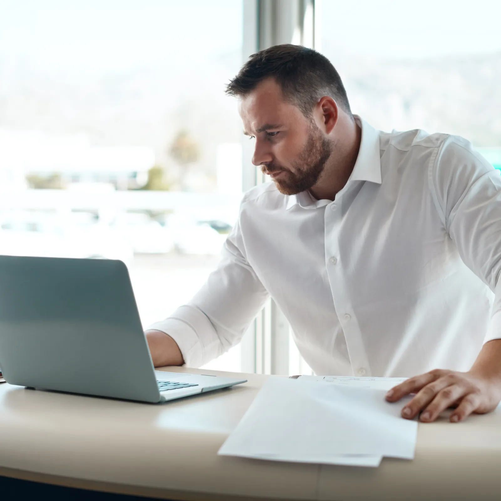 Man in white shirt working on laptop at desk with coffee cup and papers near large window.