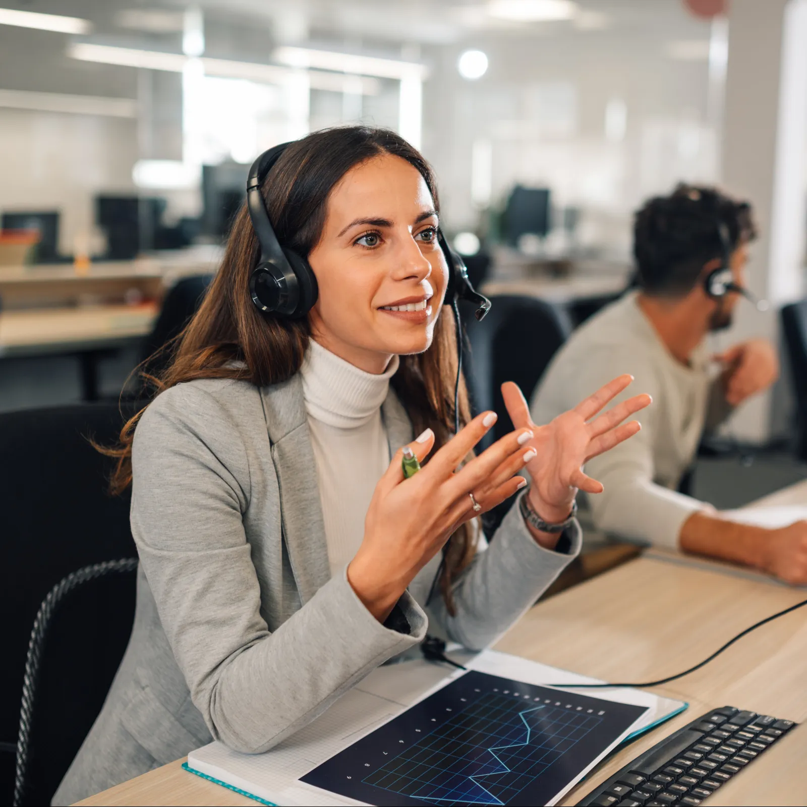 Female call center agent wearing headset smiling and gesturing while working at a computer in open office.