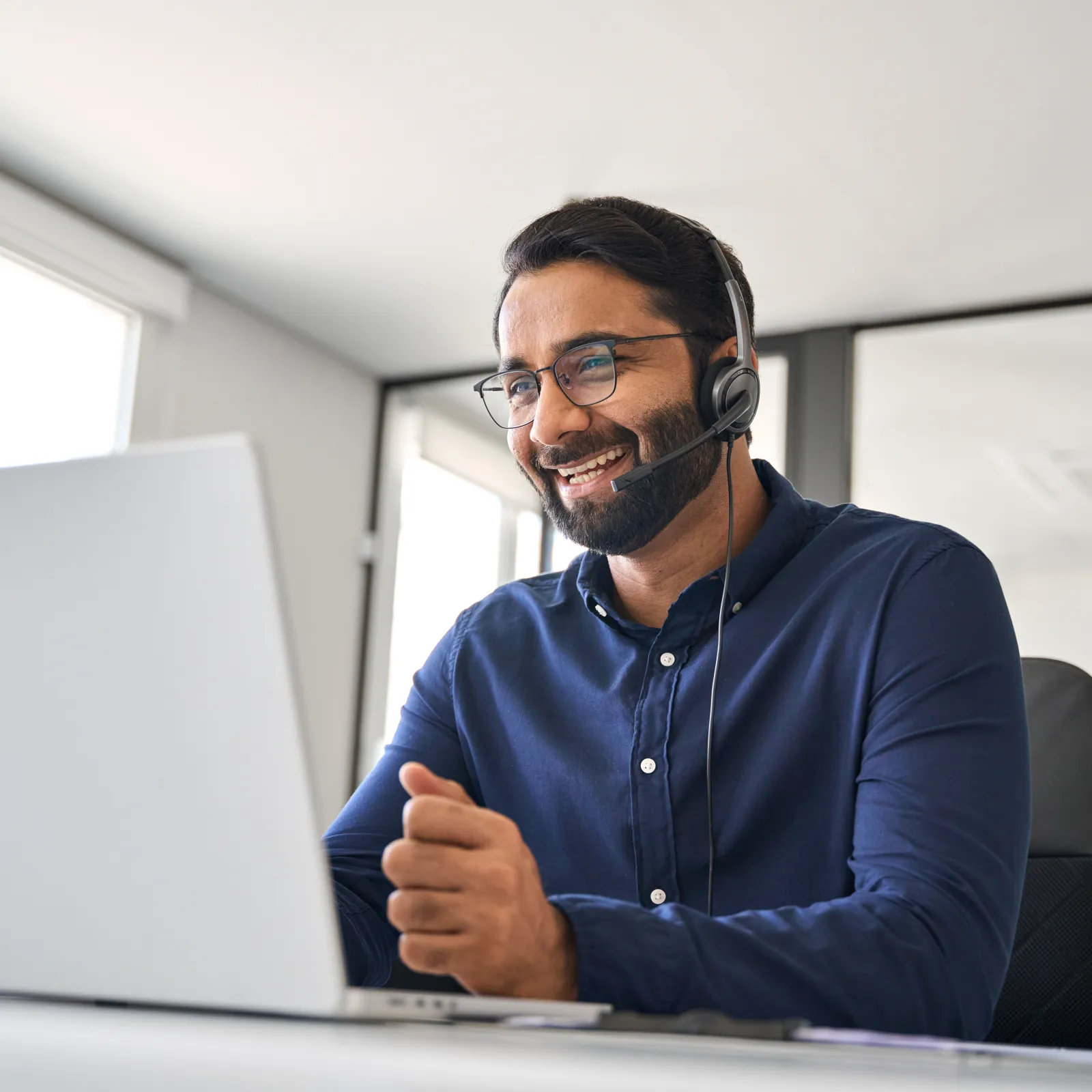 Smiling man wearing headset and glasses working on laptop in a bright modern office environment.