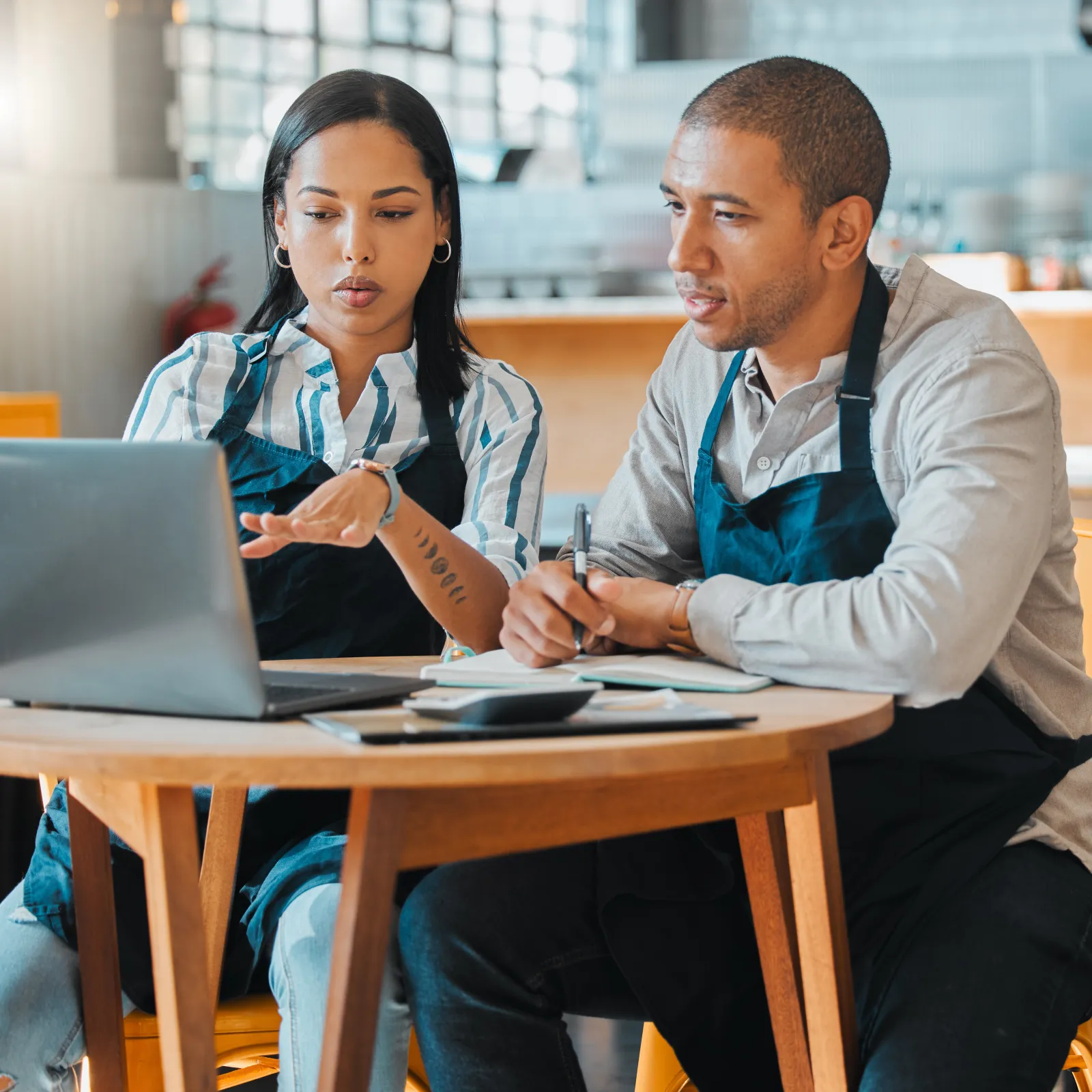 Two cafe workers in aprons discuss business plans using a laptop and notepad inside a modern coffee shop.