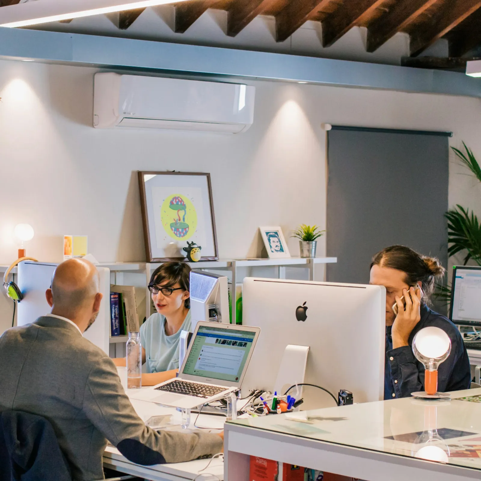 Modern office workspace with three professionals collaborating using laptops and desktop computers surrounded by plants.