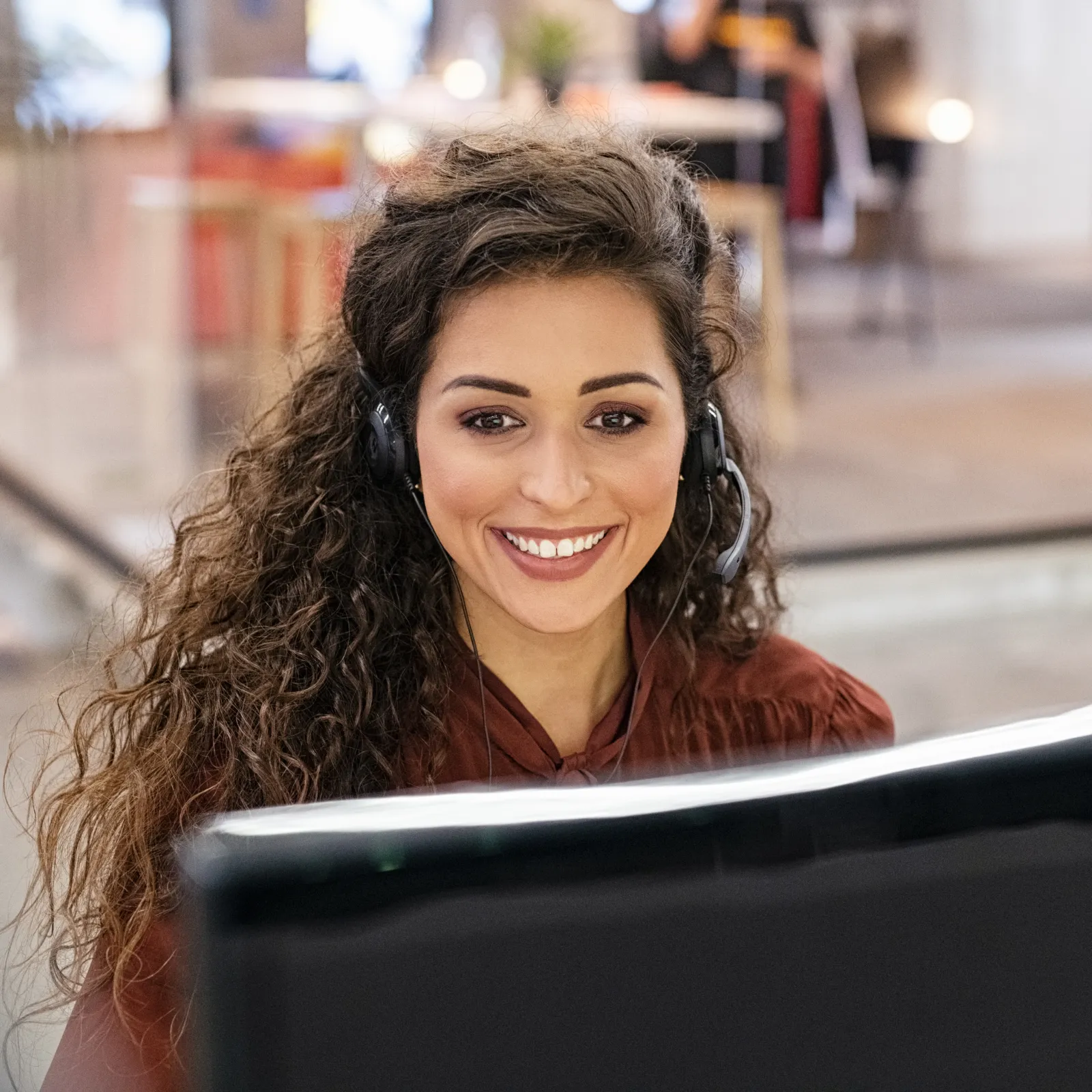 Smiling woman with headset working at computer in modern office environment with blurred background.