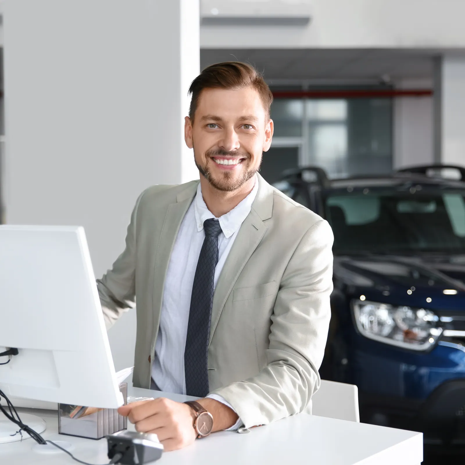 Smiling car salesman in beige suit working at desk with computer in a modern showroom with car behind him