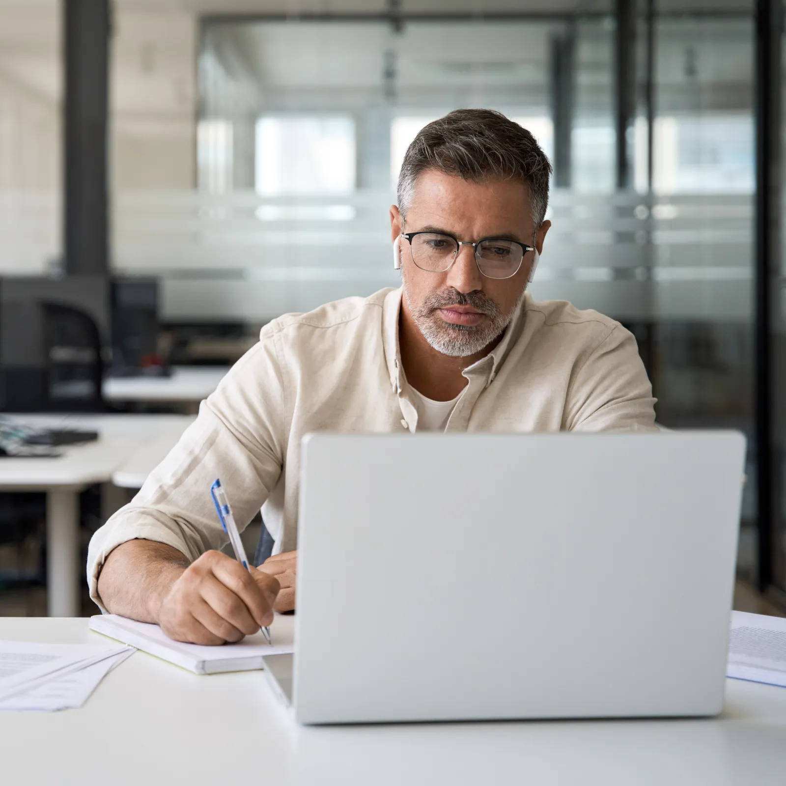 Man wearing glasses working on a laptop and taking notes in a modern office with empty desks