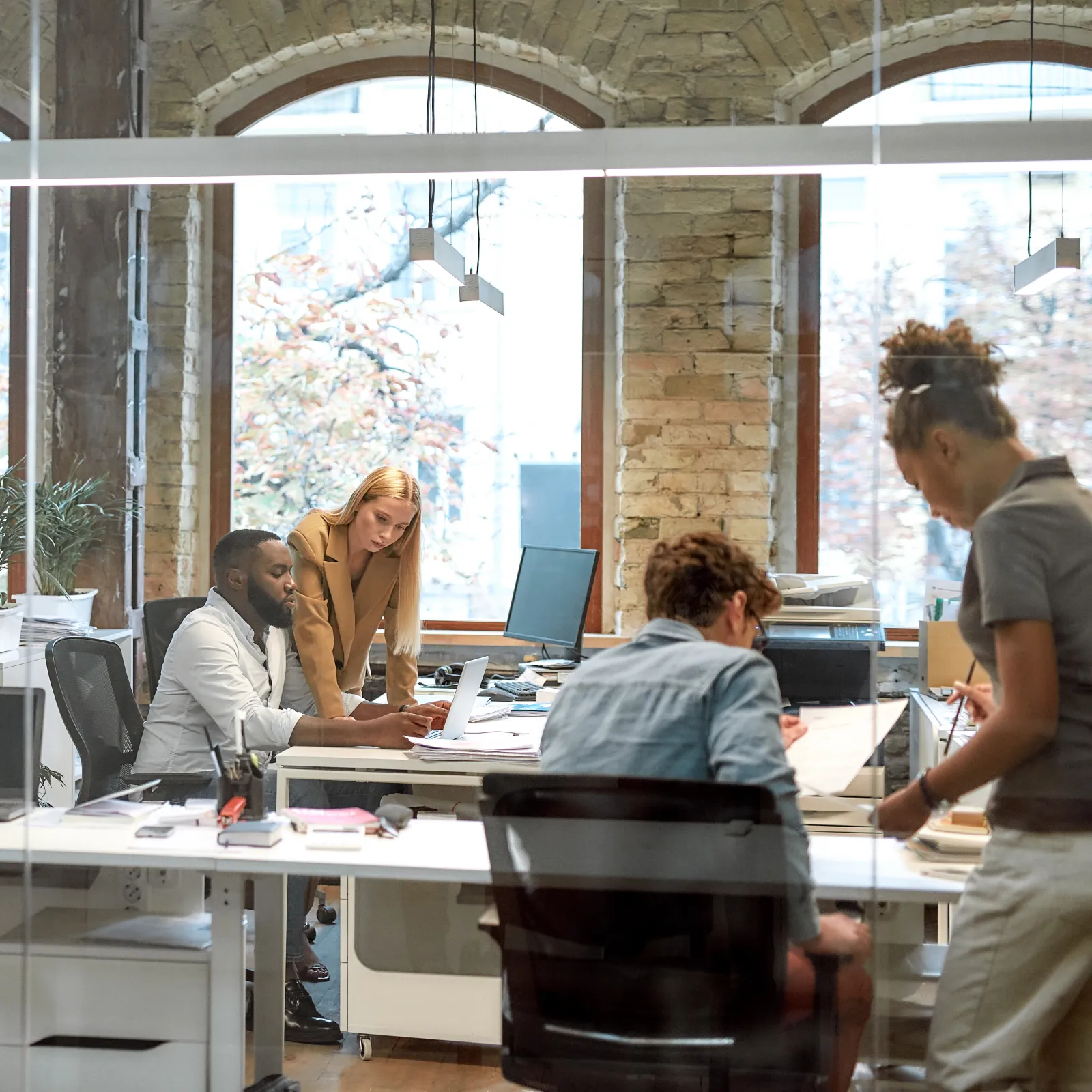 Diverse team collaborating and working on laptops in a modern office with large arched windows and exposed brick walls.