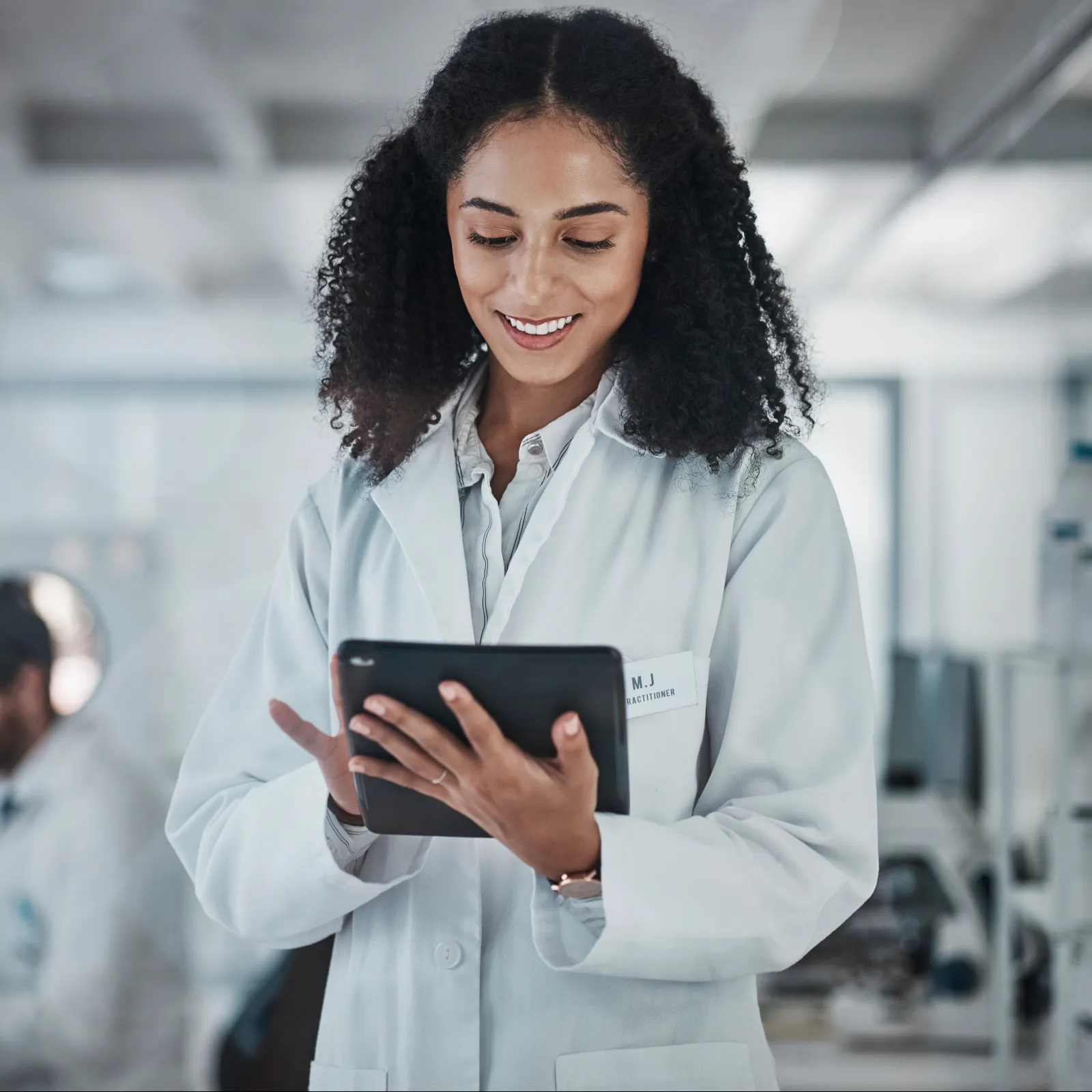 Smiling female scientist in lab coat using a tablet in a bright modern laboratory with colleague working in background