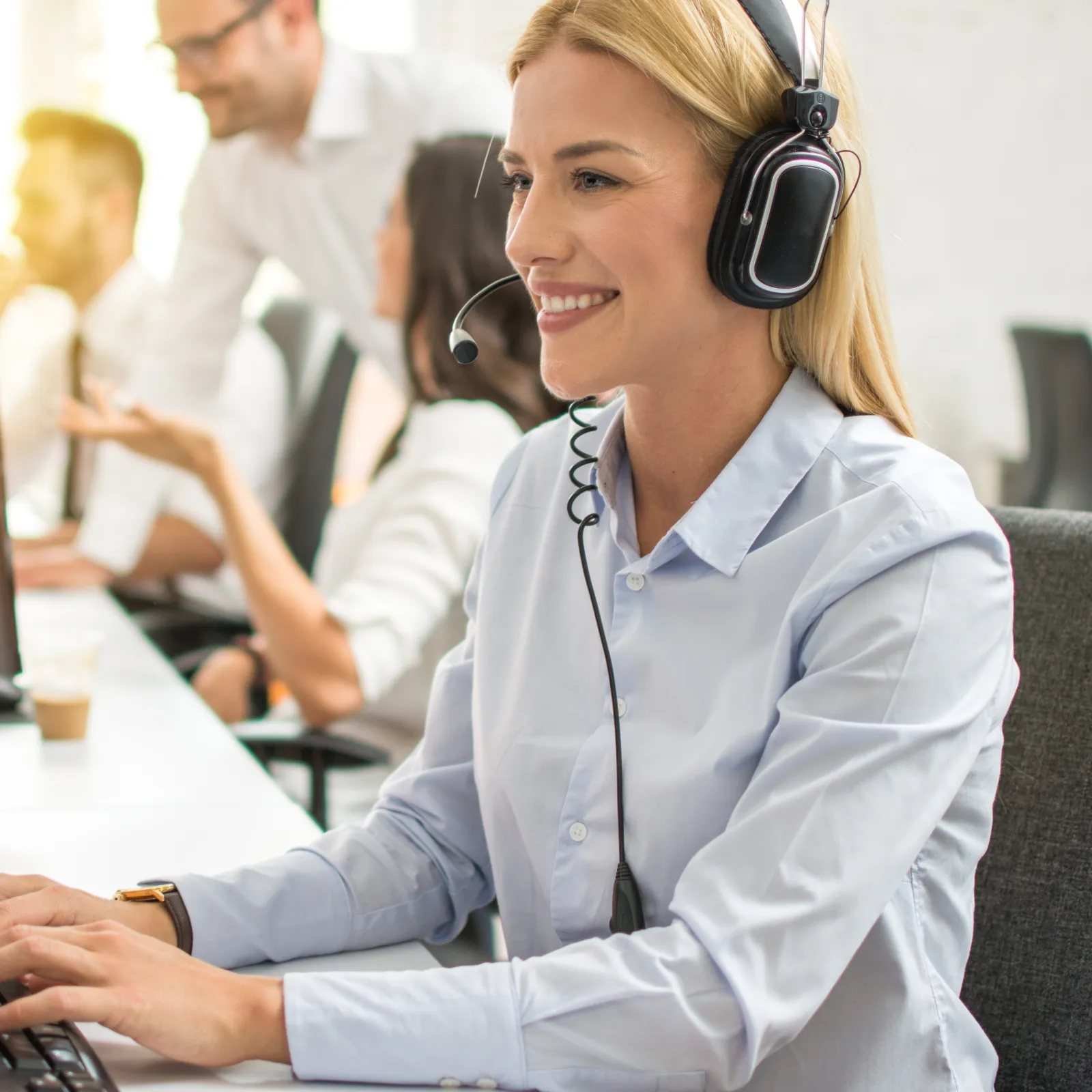 Smiling female customer service agent wearing headset typing on keyboard in busy call center with colleagues