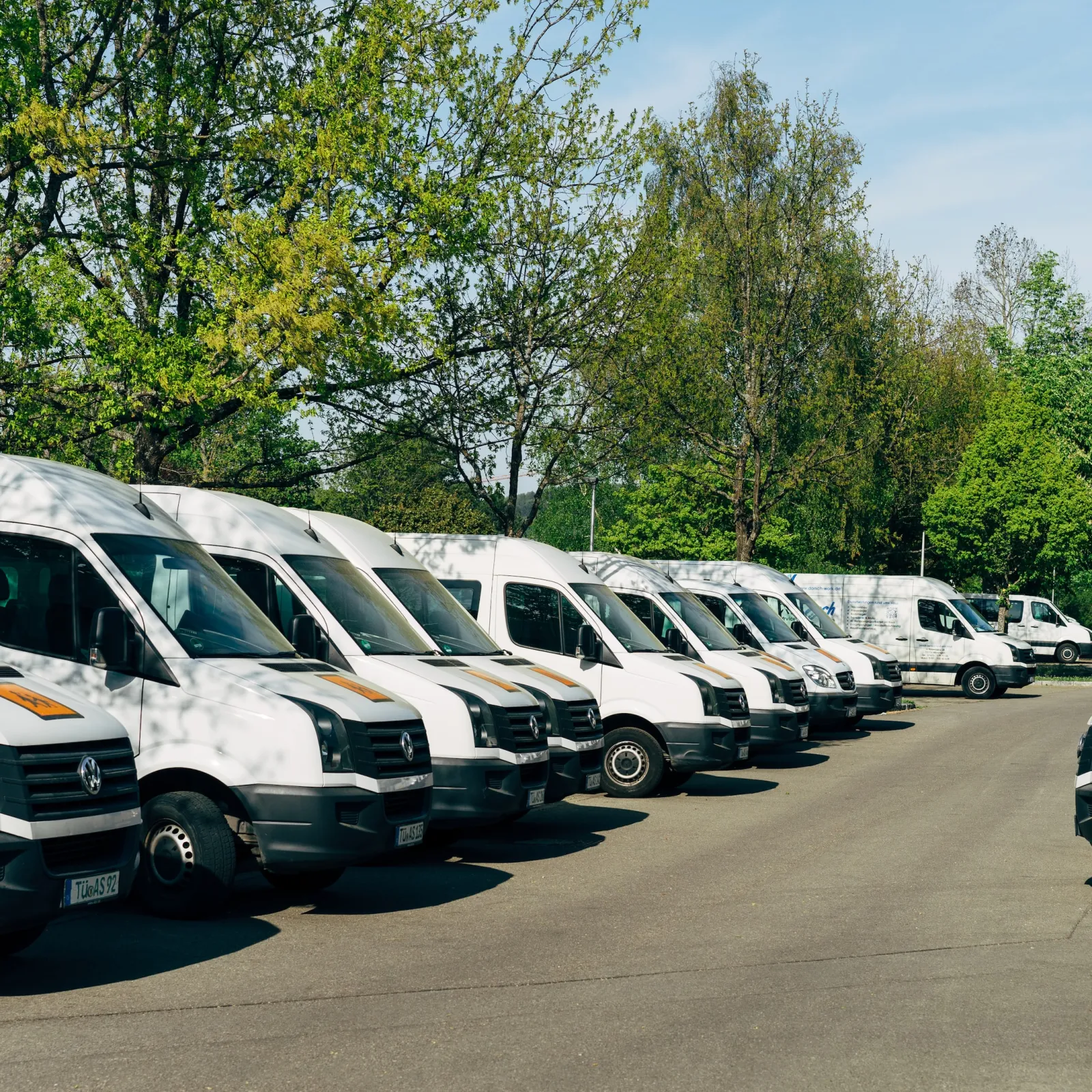Row of white delivery vans parked outdoors surrounded by green trees under a clear sky.