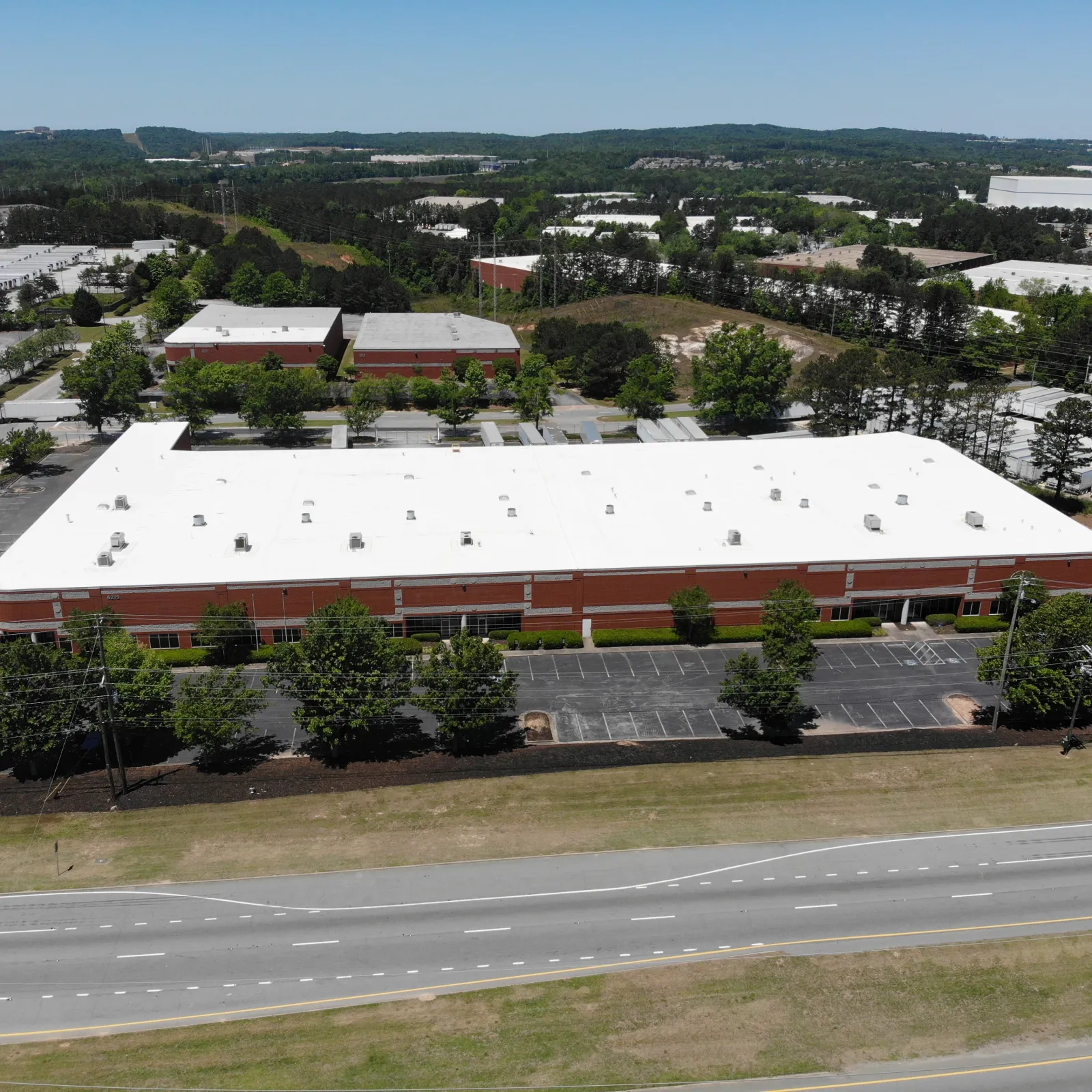 Aerial view of a large commercial warehouse with a white roof and surrounding parking lots next to a multi-lane road.