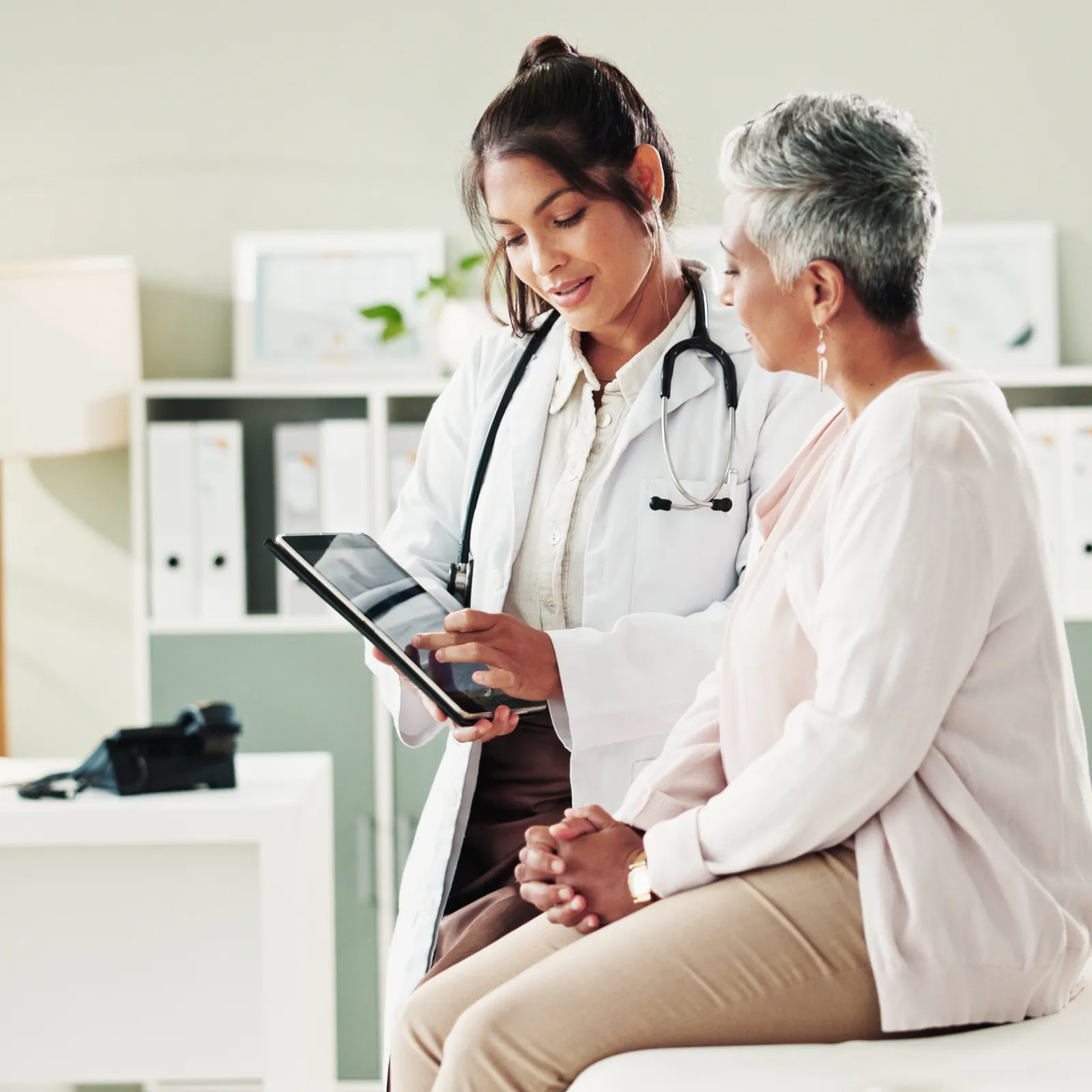 Female doctor consulting senior woman patient and showing health information on digital tablet in clinic