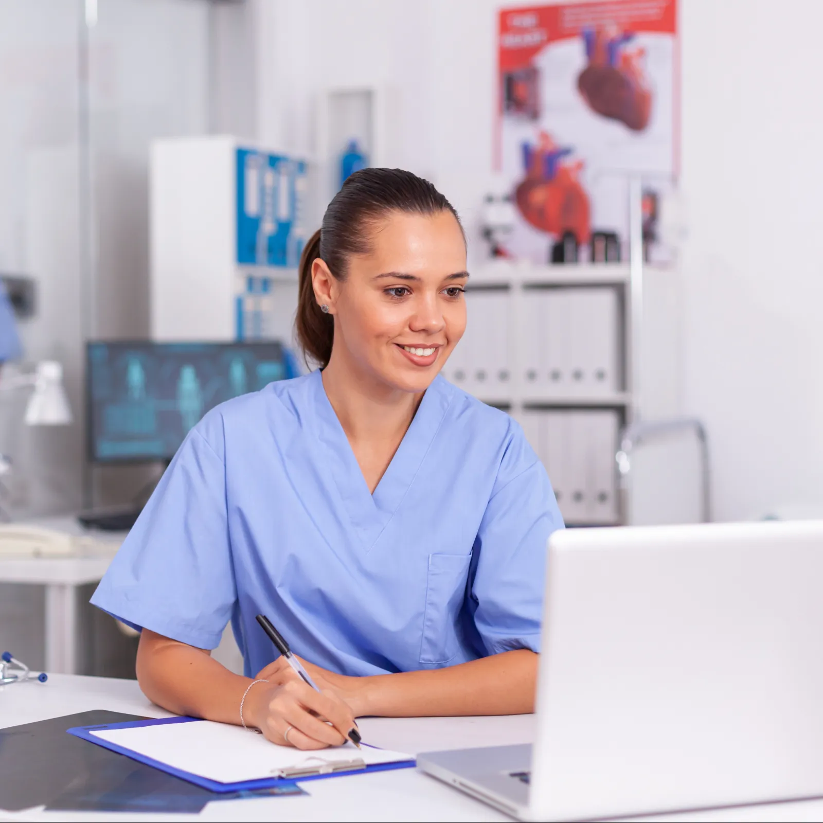 Female nurse in blue scrubs smiling while writing on clipboard and using laptop in medical office