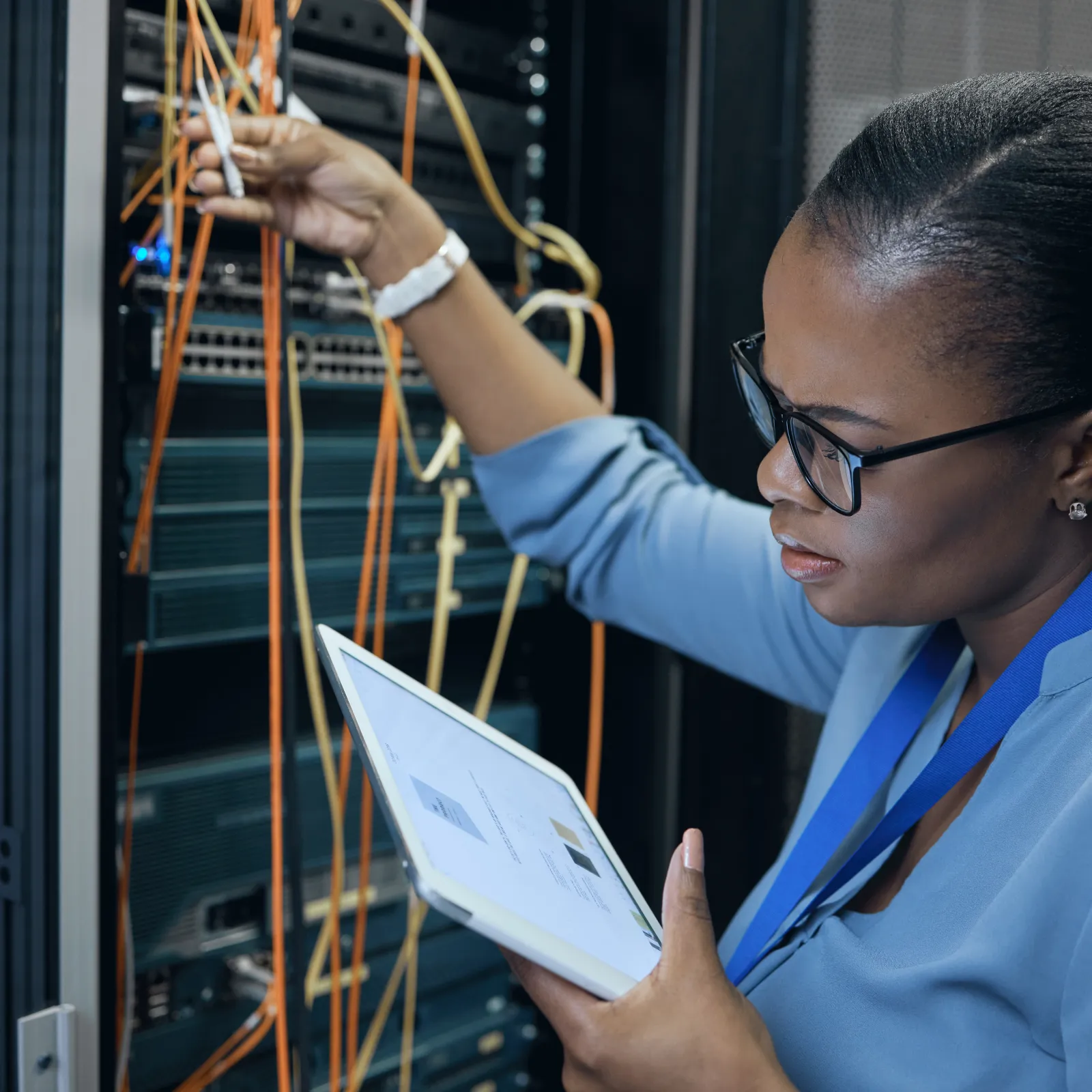 Female IT technician managing server cables while using a tablet in a data center rack room environment.