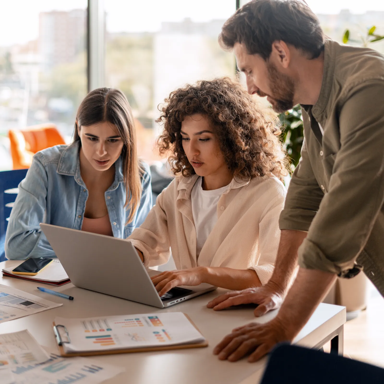 Three colleagues collaborating on a laptop with charts and documents on a bright office table.