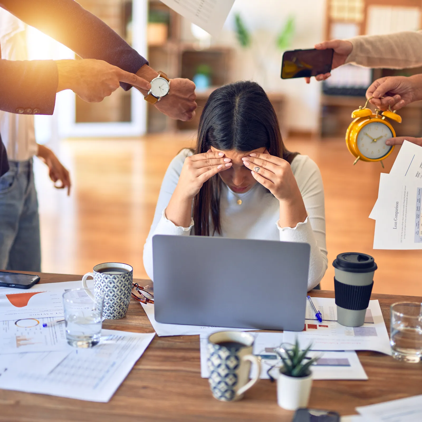 Stressed woman at desk surrounded by hands pointing, showing charts, holding clock and phone in busy office environment